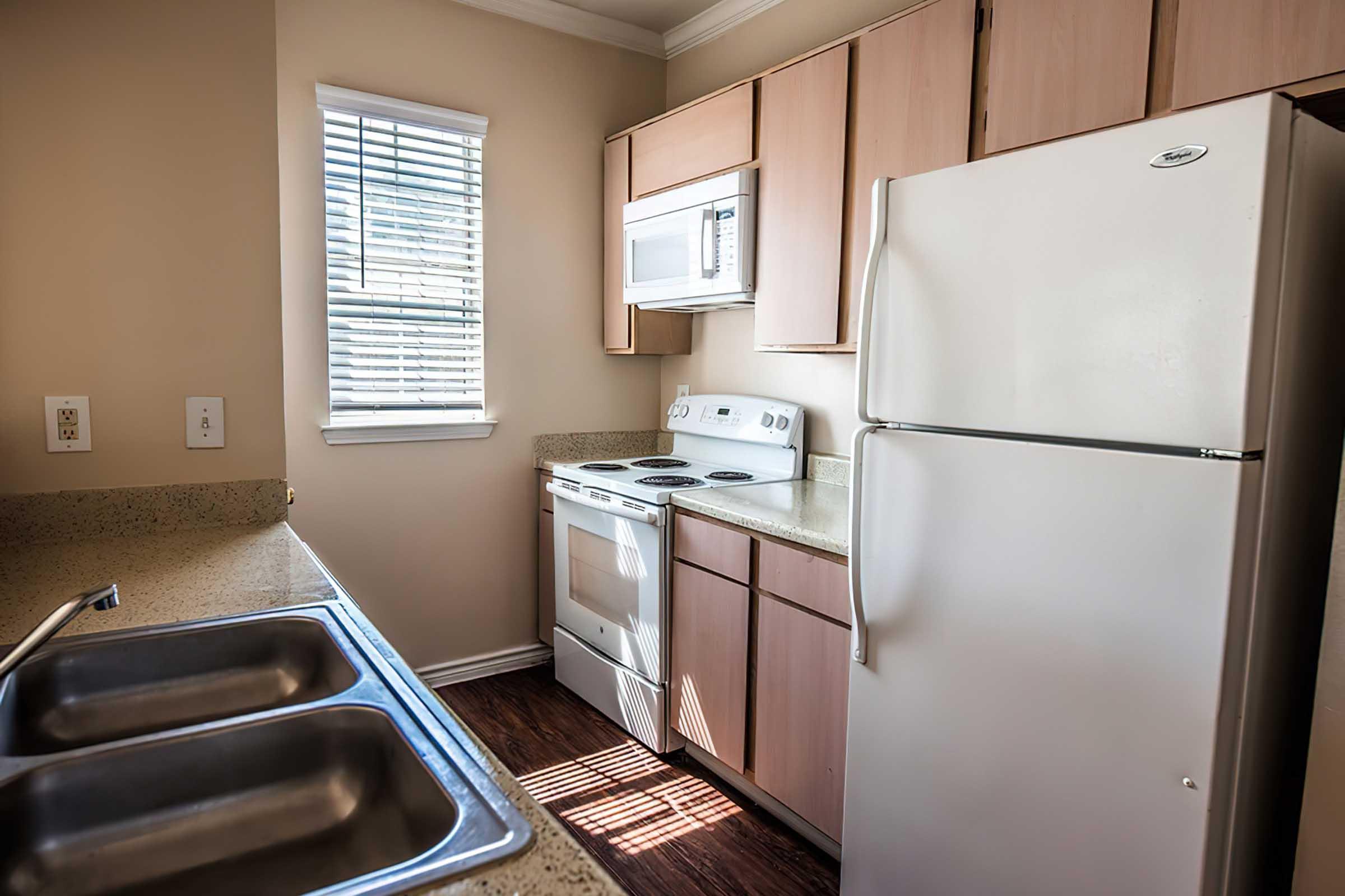 Modern kitchen featuring light wood cabinetry, white appliances including a refrigerator, stove, and microwave, granite countertops, and a double sink. Natural light comes in through a window with blinds, creating a bright and inviting atmosphere.