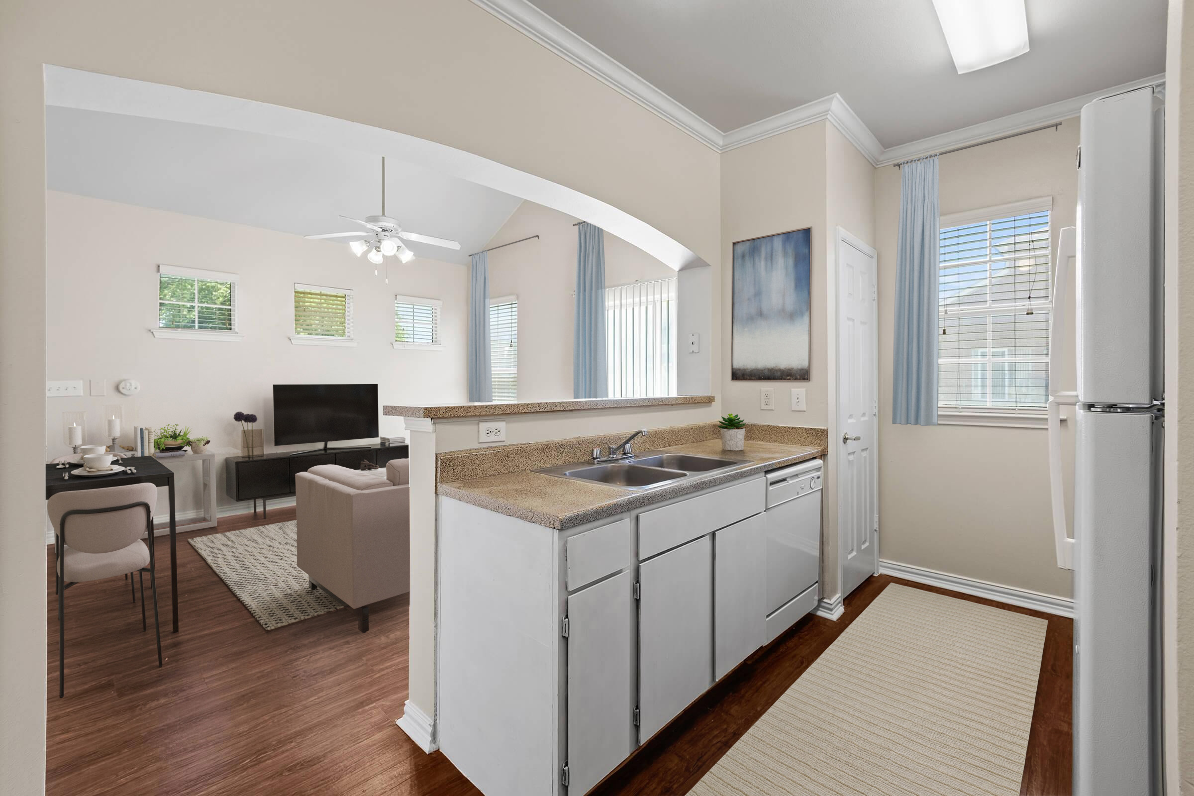 A modern kitchen with white cabinets and a double sink, opening into a cozy living area. The space features a small dining table, a sofa, and a TV. Large windows let in natural light, and there are decorative elements like a rug and plants, creating a warm and inviting atmosphere.