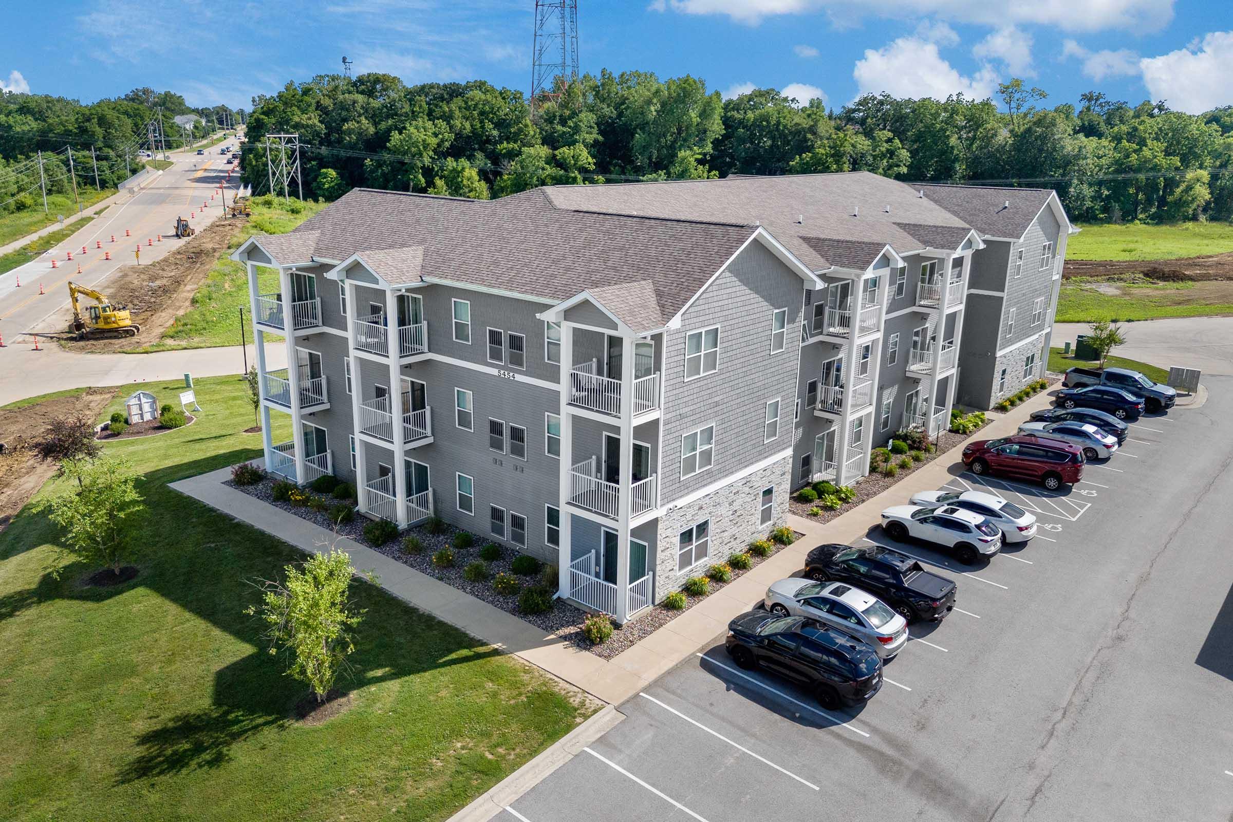 A modern multi-unit apartment building with gray siding and balconies, surrounded by well-maintained landscaping. Several parked cars are in the foreground, while construction and power lines are visible in the background. Bright blue sky and trees add to the setting.