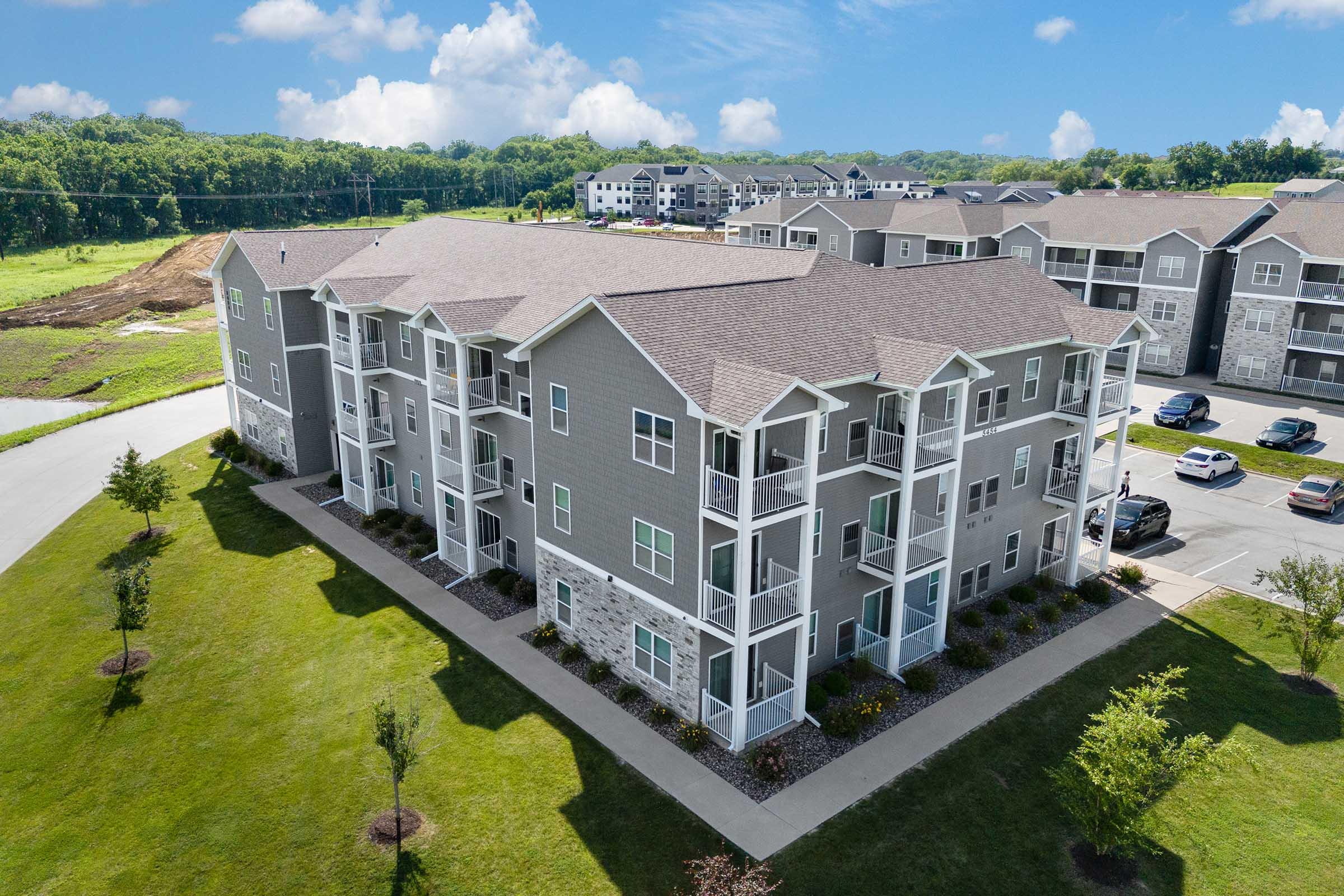 Aerial view of a modern three-story apartment building featuring a light gray exterior with stone accents. Surrounding green landscaping includes grass and small trees. In the background, additional apartment buildings can be seen under a sunny sky with scattered clouds.