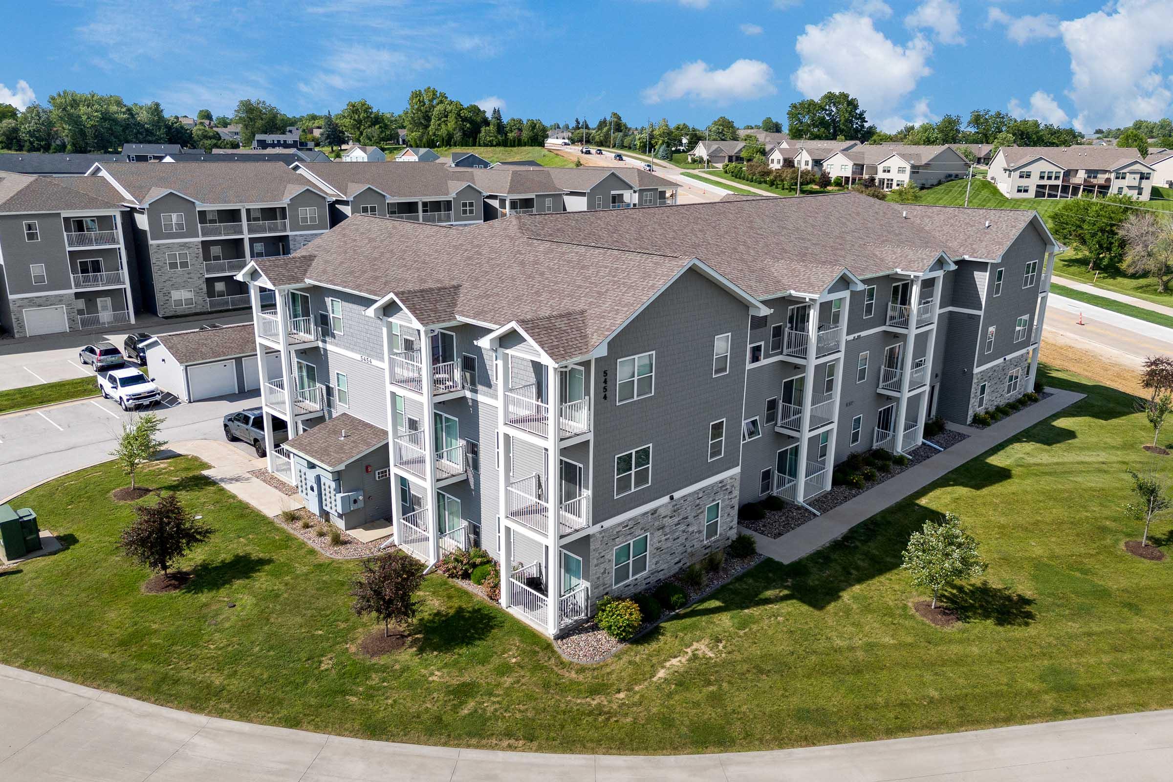 Aerial view of a modern multi-unit apartment building with gray siding and stone accents. Surrounding green space includes trees and landscaped areas. Driveway and additional apartment buildings are visible in the background, under a partly cloudy blue sky.
