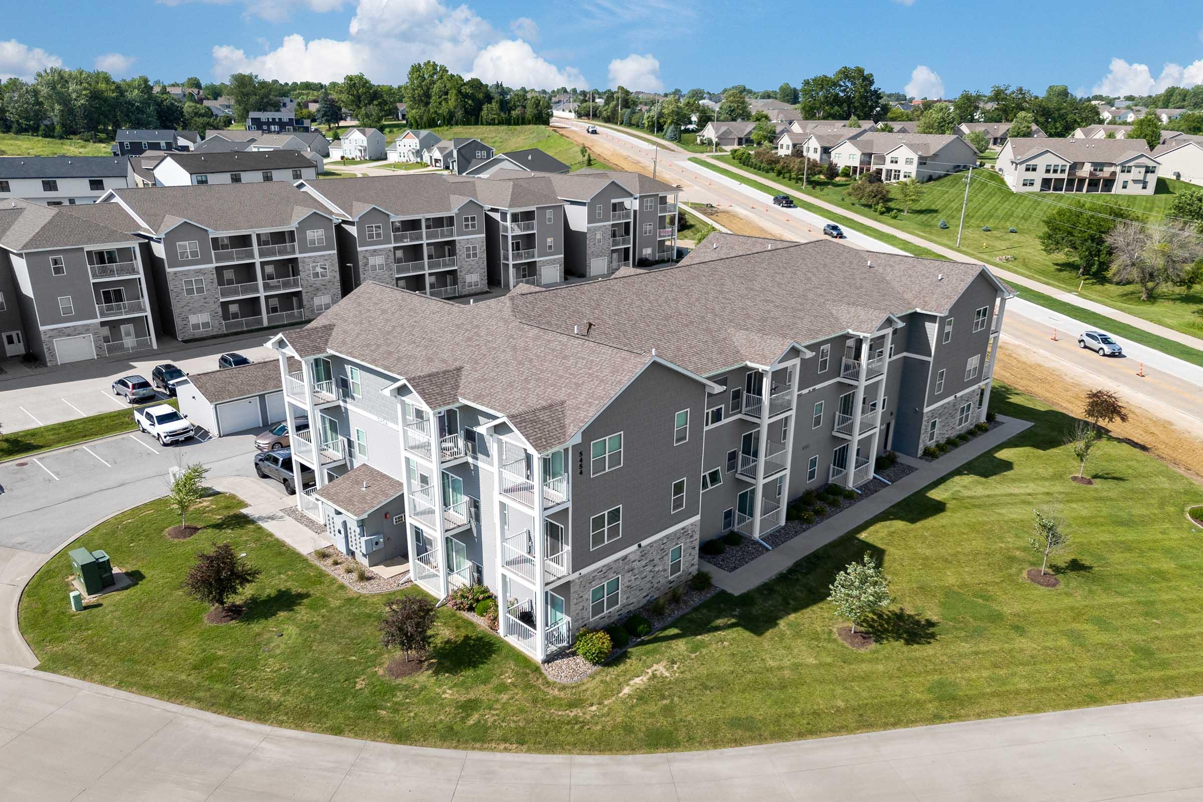Aerial view of a modern apartment complex with multiple gray buildings surrounded by green lawns, trees, and parking spaces. In the background, more residential buildings and a road can be seen under a clear blue sky with scattered clouds.