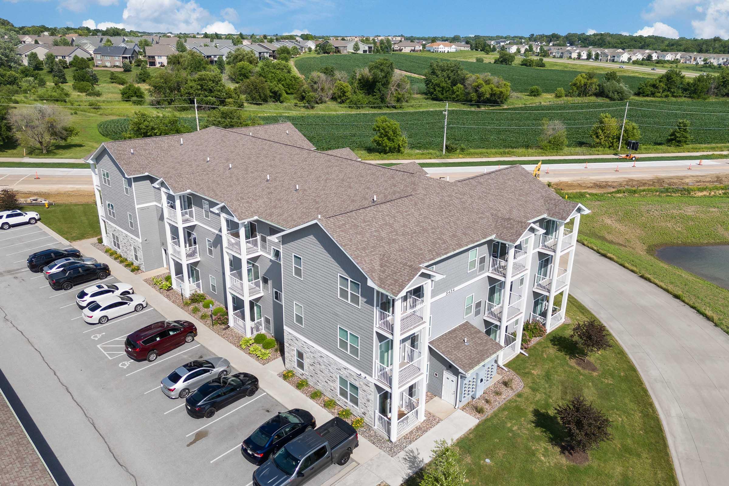 Aerial view of a three-story residential apartment building with a modern design, featuring balconies and a landscaped parking area in front. Surrounding the building are green fields and trees, with a road nearby. Clear blue skies and fluffy clouds complete the scenic backdrop.