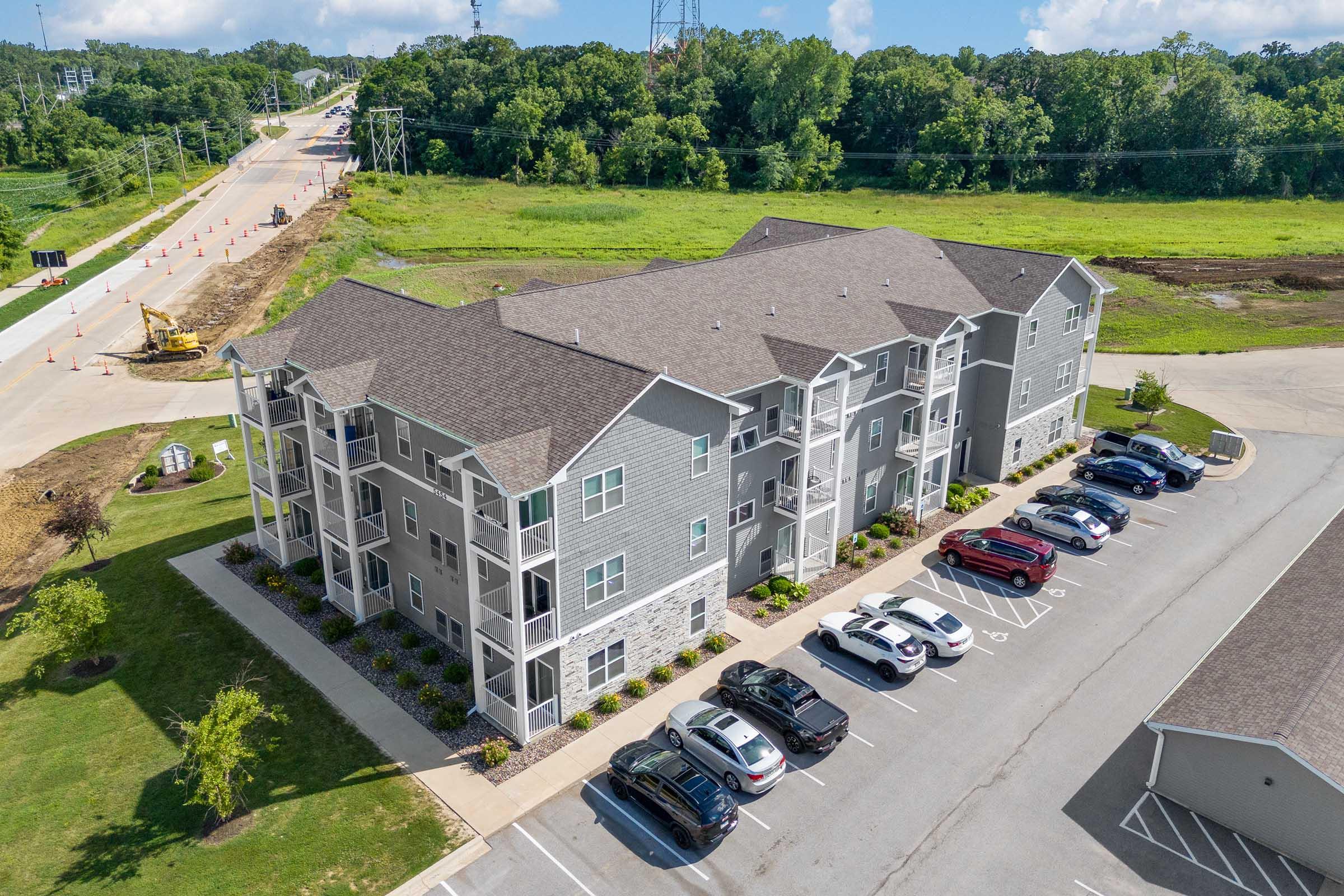 Aerial view of a modern apartment complex featuring several three-story buildings with gray siding and stone accents. In the foreground, there is a parking lot with various cars. Surrounding greenery and a road under maintenance can be seen in the background. Clear blue sky with some clouds overhead.