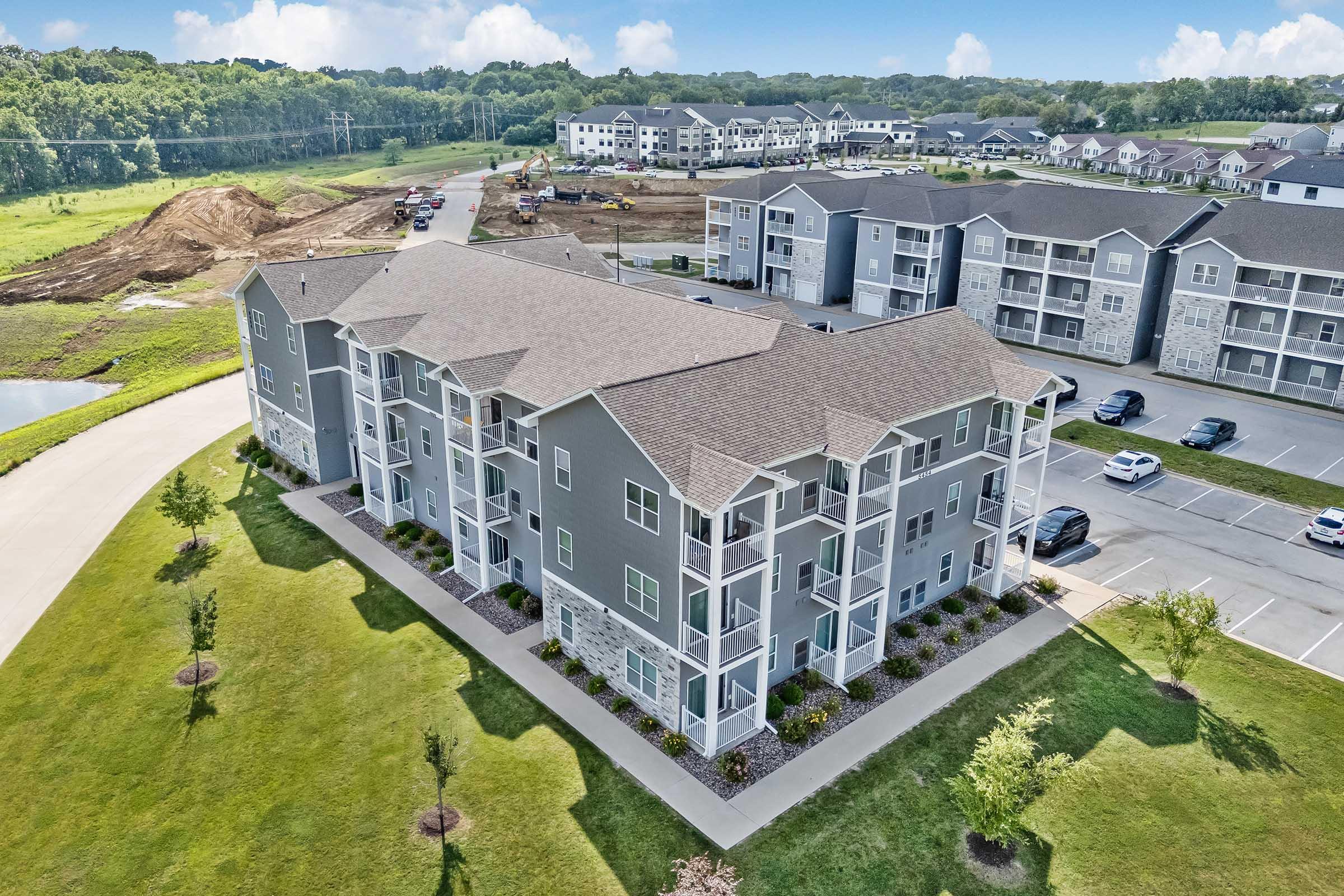 Aerial view of a modern apartment complex featuring several three-story buildings. The foreground shows a well-manicured lawn with newly planted trees, while in the background, additional residential buildings are visible. A construction site and a winding road are also present, indicating ongoing development.