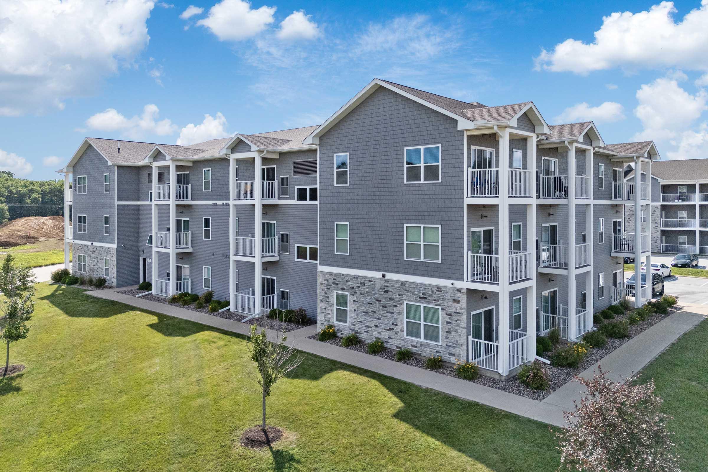 A modern three-story apartment building featuring gray siding and stone accents. There are balconies on several units, well-maintained lawns with small trees and shrubs surrounding the building. The sky is bright blue with scattered clouds, indicating a sunny day.
