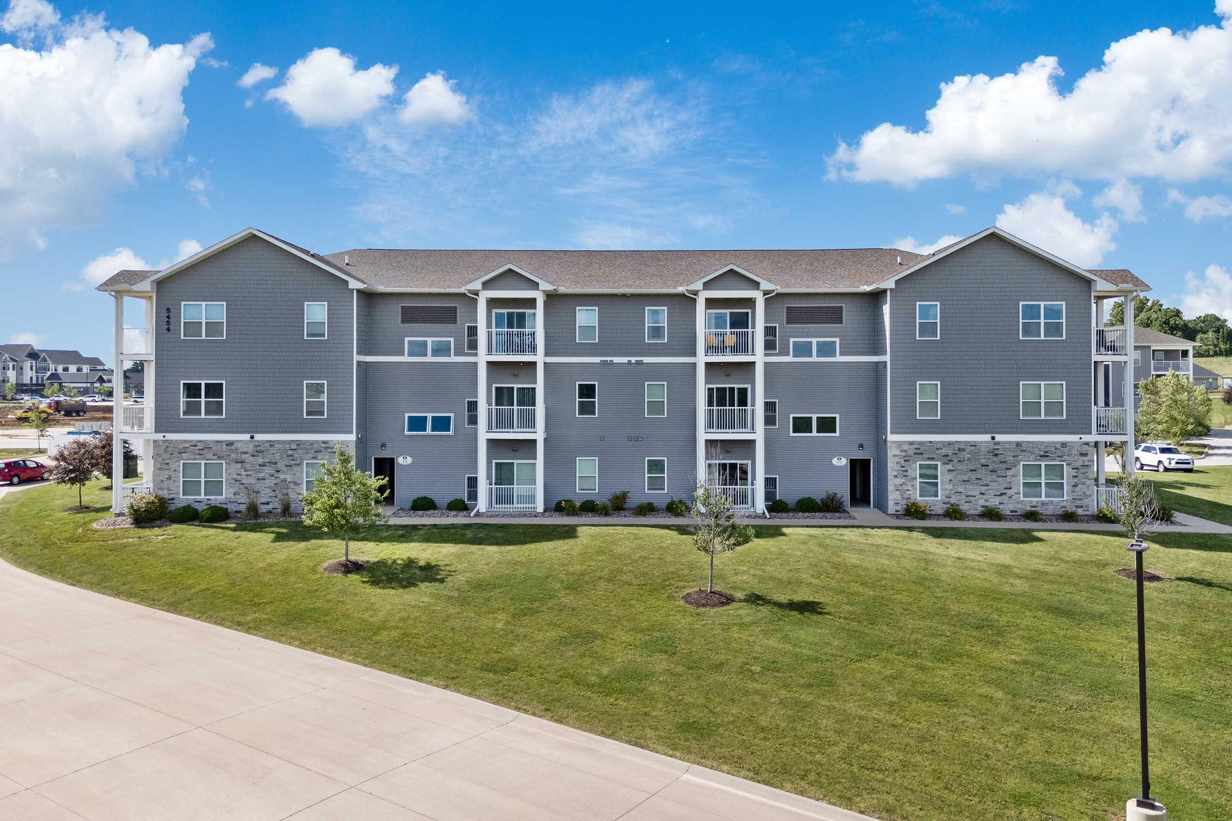A three-story apartment building with gray siding, multiple balconies, and landscaped greenery in front. The building features several windows and a concrete pathway leading to it, under a clear blue sky with fluffy clouds. Parking areas are visible in the background.