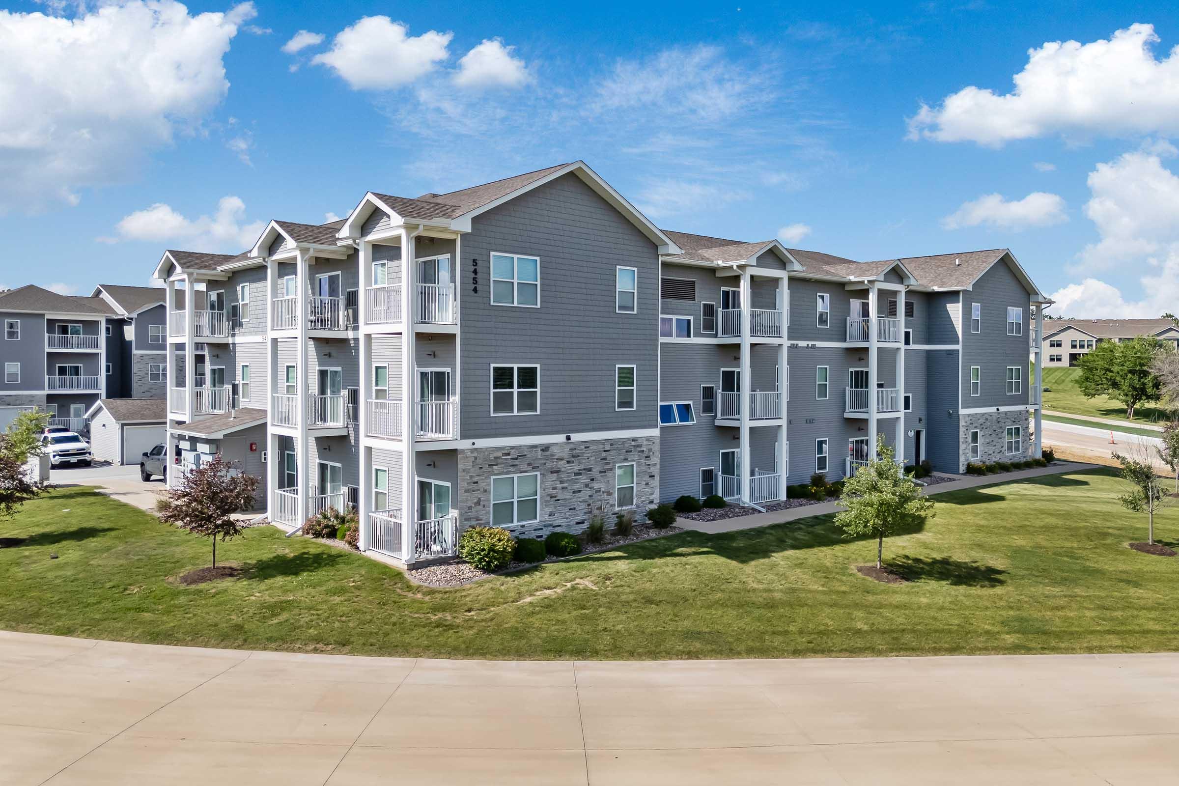 A modern apartment building with multiple stories, featuring a combination of stone and grey siding. The structure has balconies and landscaped areas with trees and grass in the foreground, under a blue sky with scattered clouds. A paved road curves in front of the building.
