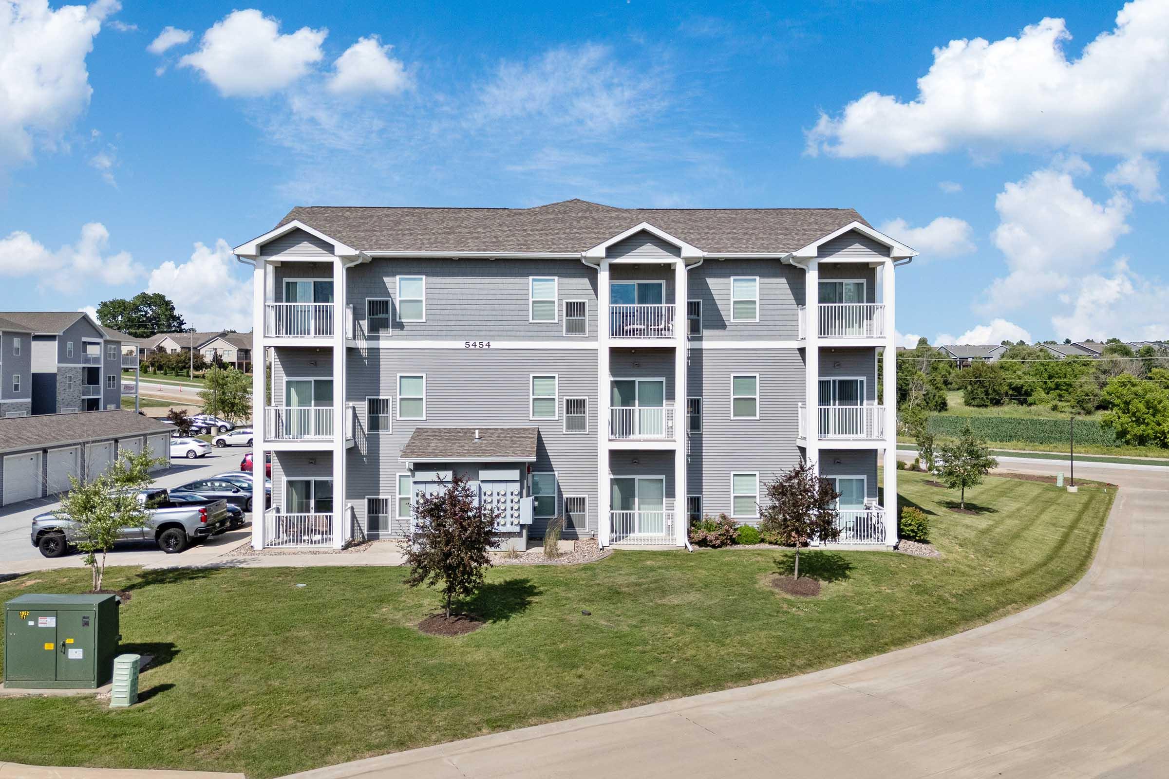 Multistory residential building with a gray exterior, featuring balconies on each unit. Surrounded by landscaped grounds, a paved driveway leads up to the building. The sky is bright blue with fluffy clouds, and there are green fields in the background.