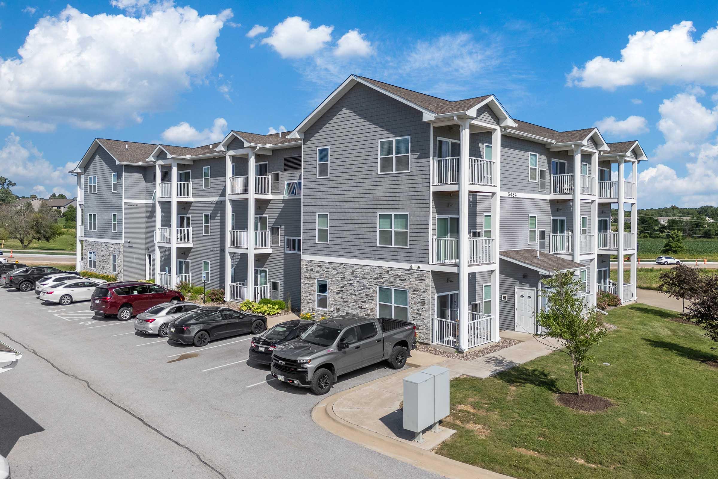 A modern three-story apartment building with a mix of gray siding and stone accents. The building features balconies and multiple parking spaces in front. Lush greenery surrounds the area under a bright blue sky with scattered clouds.