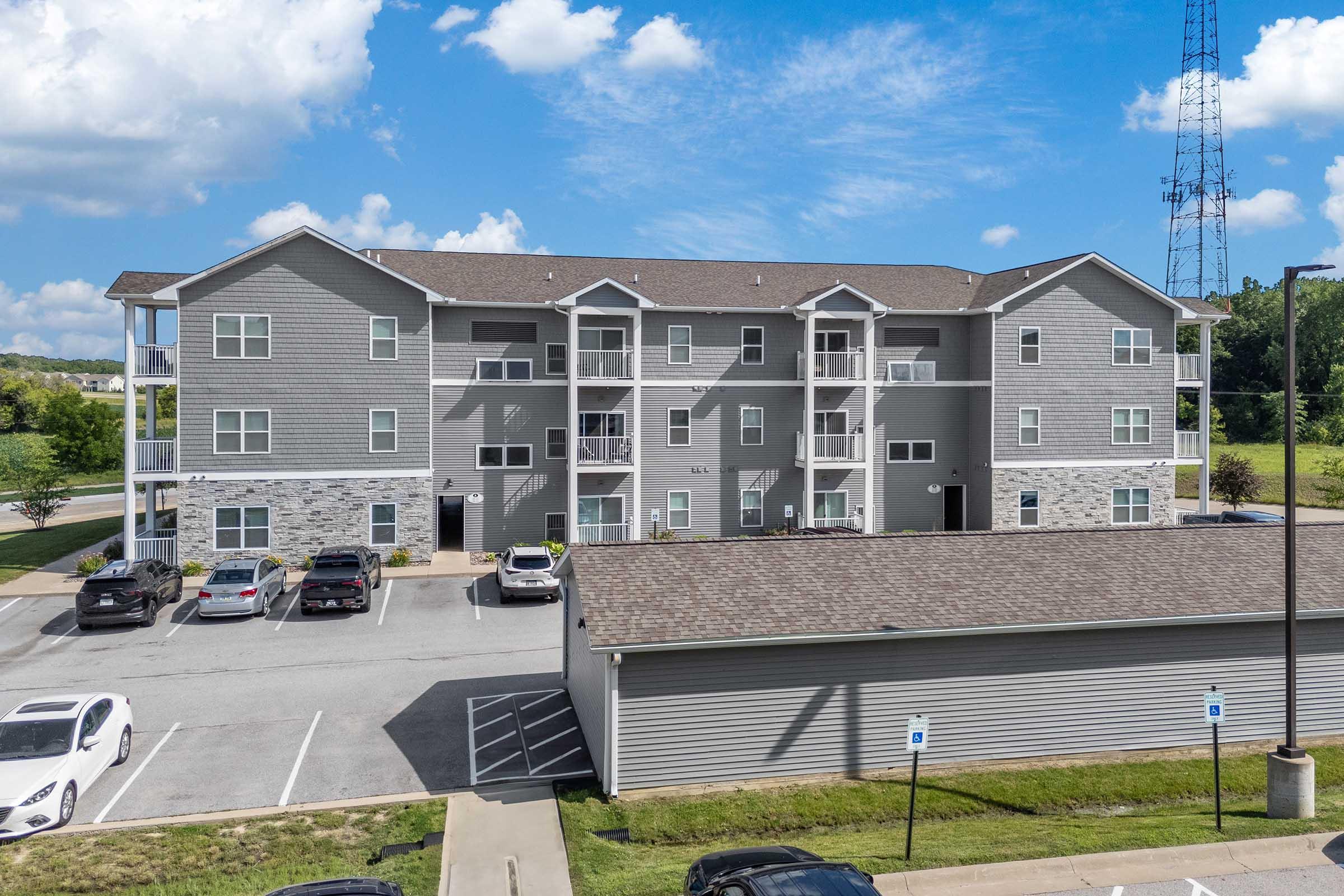 A multi-unit residential building with a gray exterior featuring a mix of stone accents. The building has three stories, balconies, and several parked cars in front. A communication tower is visible in the background against a blue sky with fluffy clouds.
