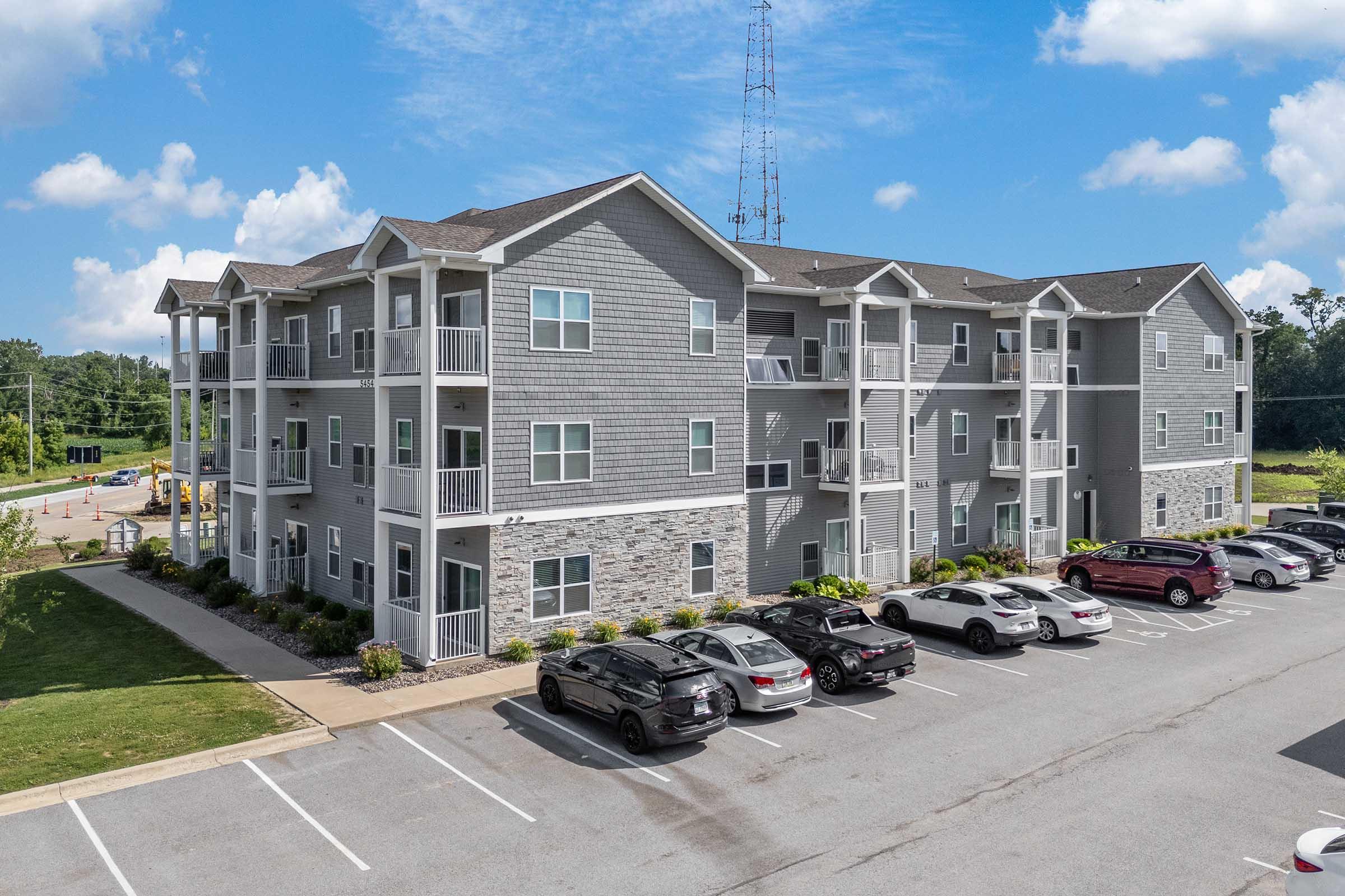 Multi-unit apartment building with a modern design featuring grey siding and stone accents. Balconies on each floor overlook a parking lot with several vehicles. The surrounding area includes green grass and a clear blue sky with some clouds.
