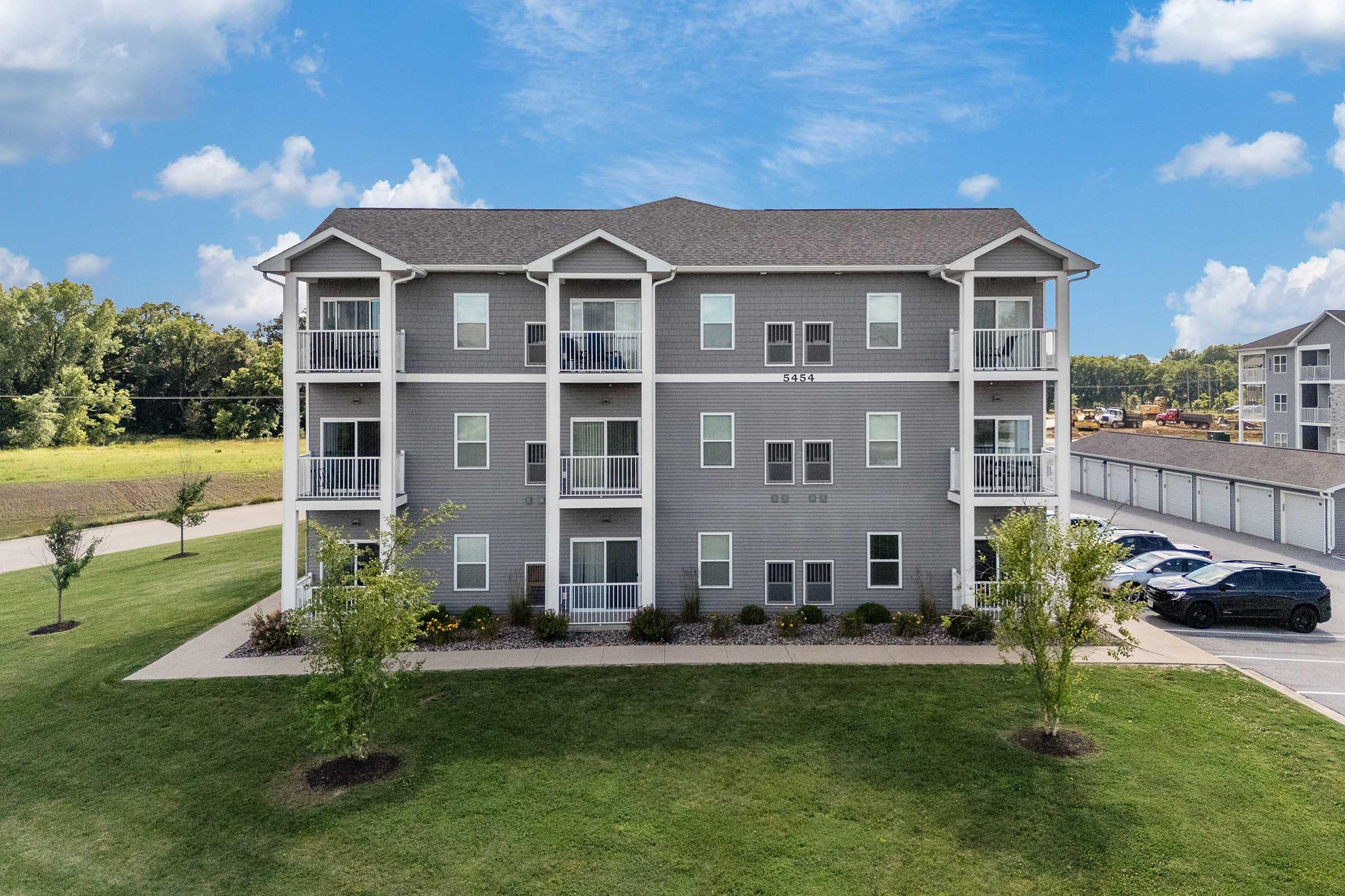 A three-story apartment building with a gray exterior and white trim. It features multiple balconies and landscaped areas with small trees and flower beds. In the background, there is a clear blue sky with white clouds and a parking area with vehicles.