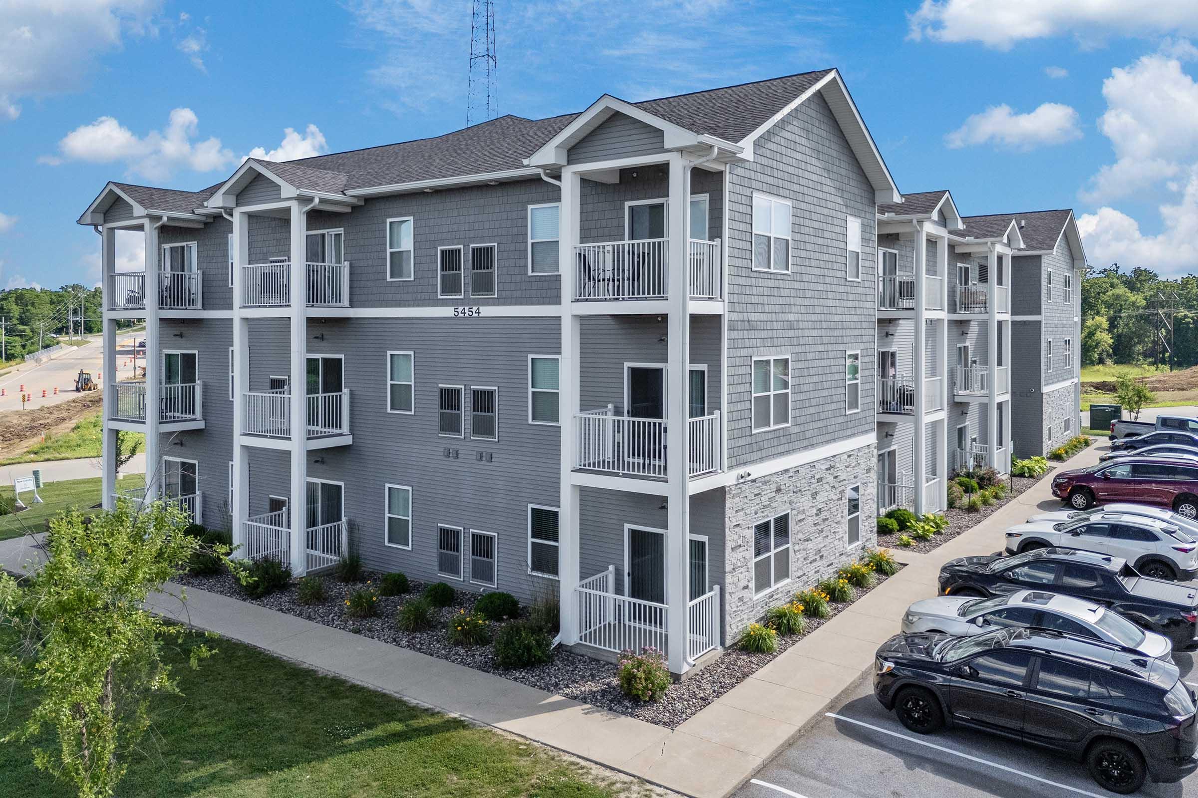 A modern multi-unit apartment building with a gray exterior, white railings, and landscaped grounds. The building features multiple balconies and parking spaces in front, with trees and grass nearby. The sky is clear and sunny, creating a bright atmosphere.