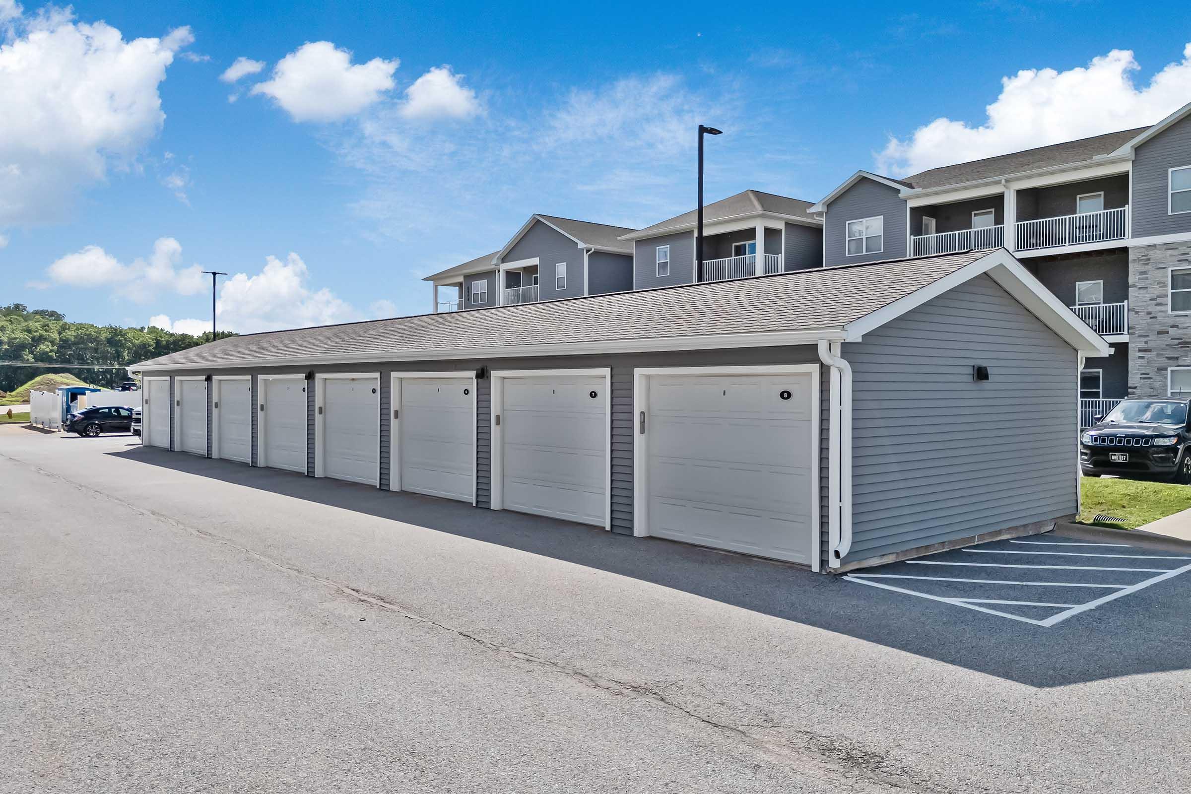 A row of gray garage doors with a parked vehicle in front, situated next to a multistory residential building. The setting is outdoors with a clear blue sky and a few clouds, illustrating a suburban environment.