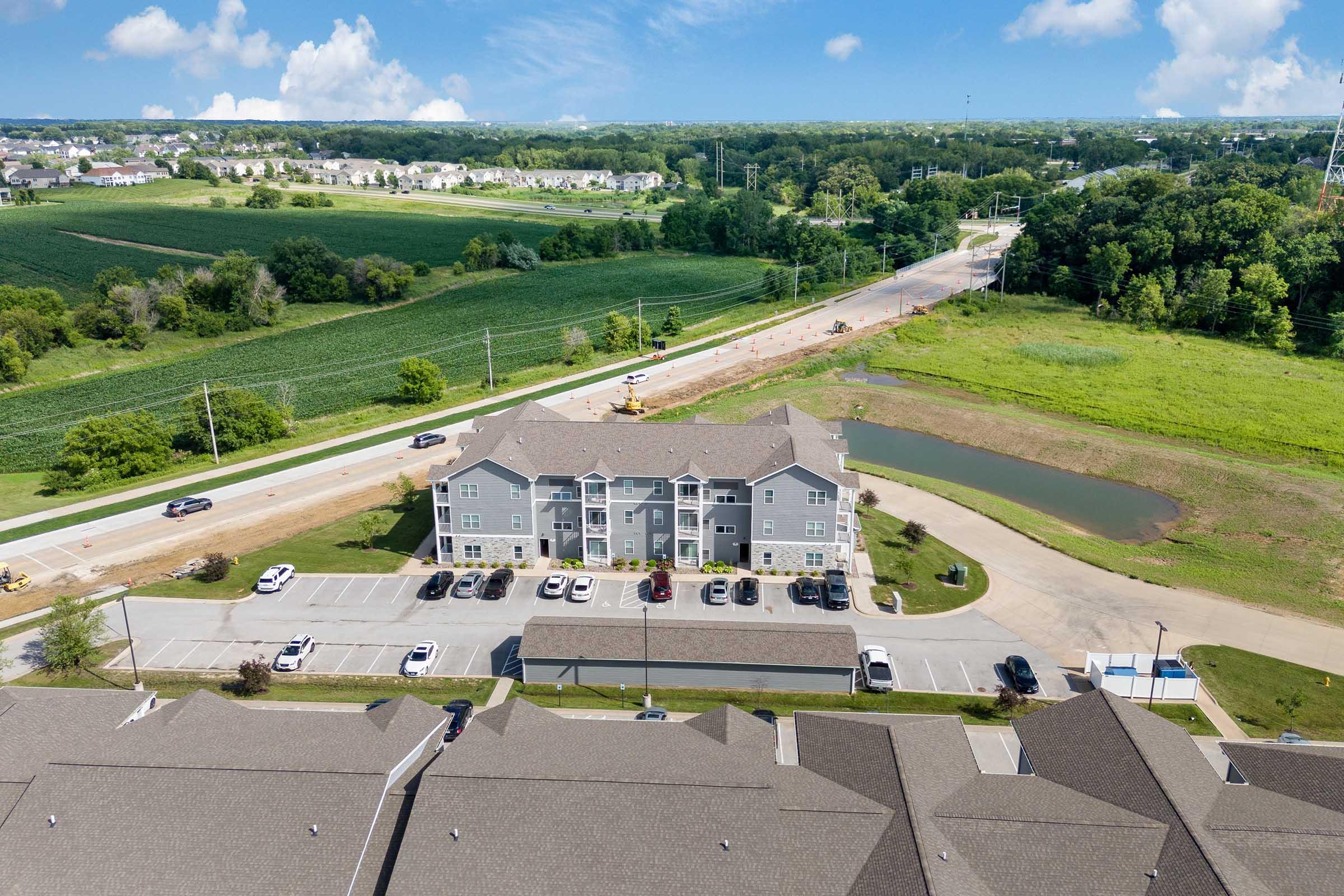 Aerial view of a residential complex with multiple buildings, surrounded by green fields and a road. Parking spaces are visible in front of the buildings, along with a small pond on the right side. Blue sky with a few clouds completes the serene landscape.