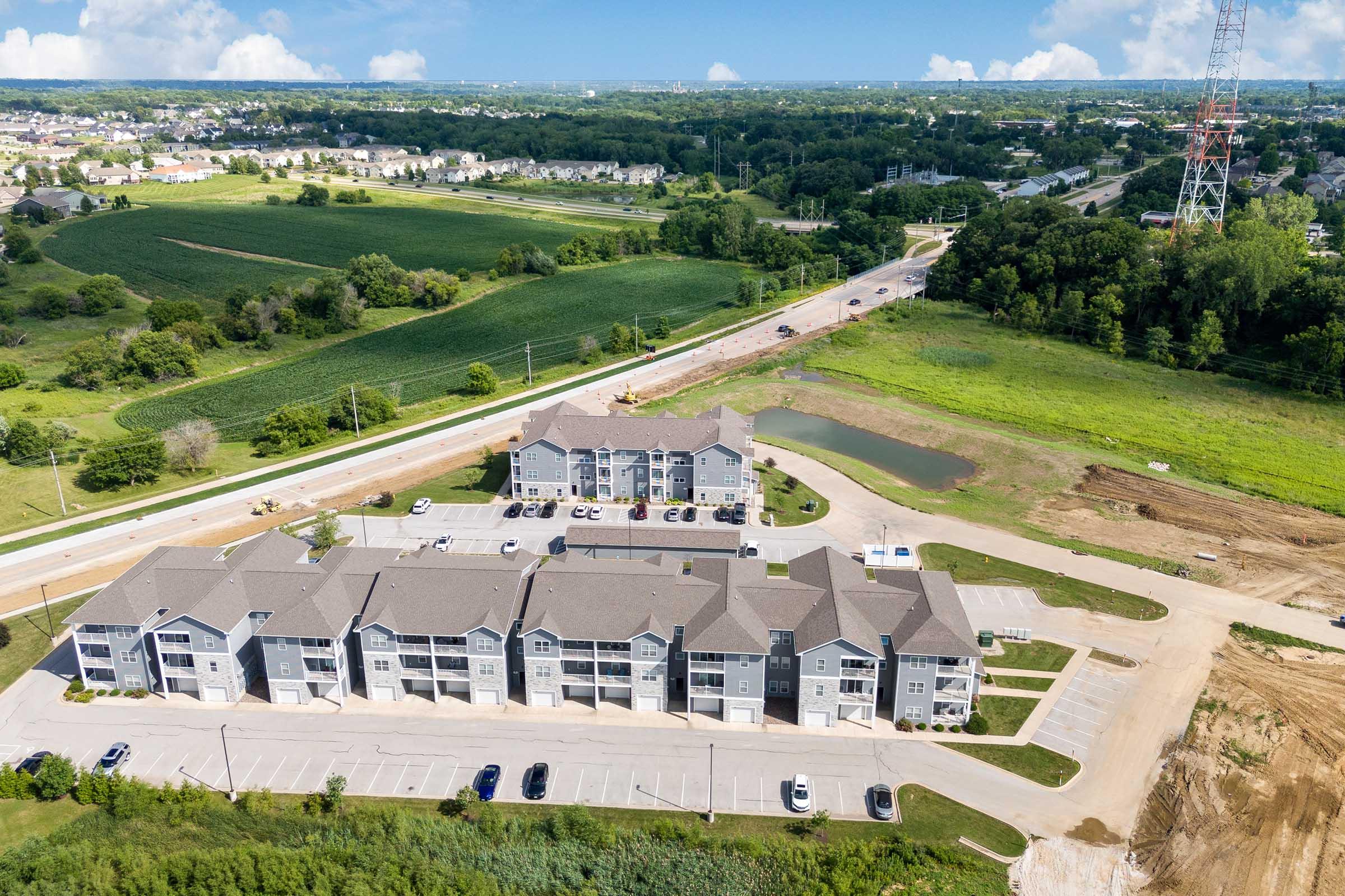 Aerial view of a residential apartment complex with multiple buildings and parking spaces. Surrounding the complex are green fields and trees, with a highway visible in the background. A small pond is situated near the buildings. The image captures a blend of urban development and natural landscape.