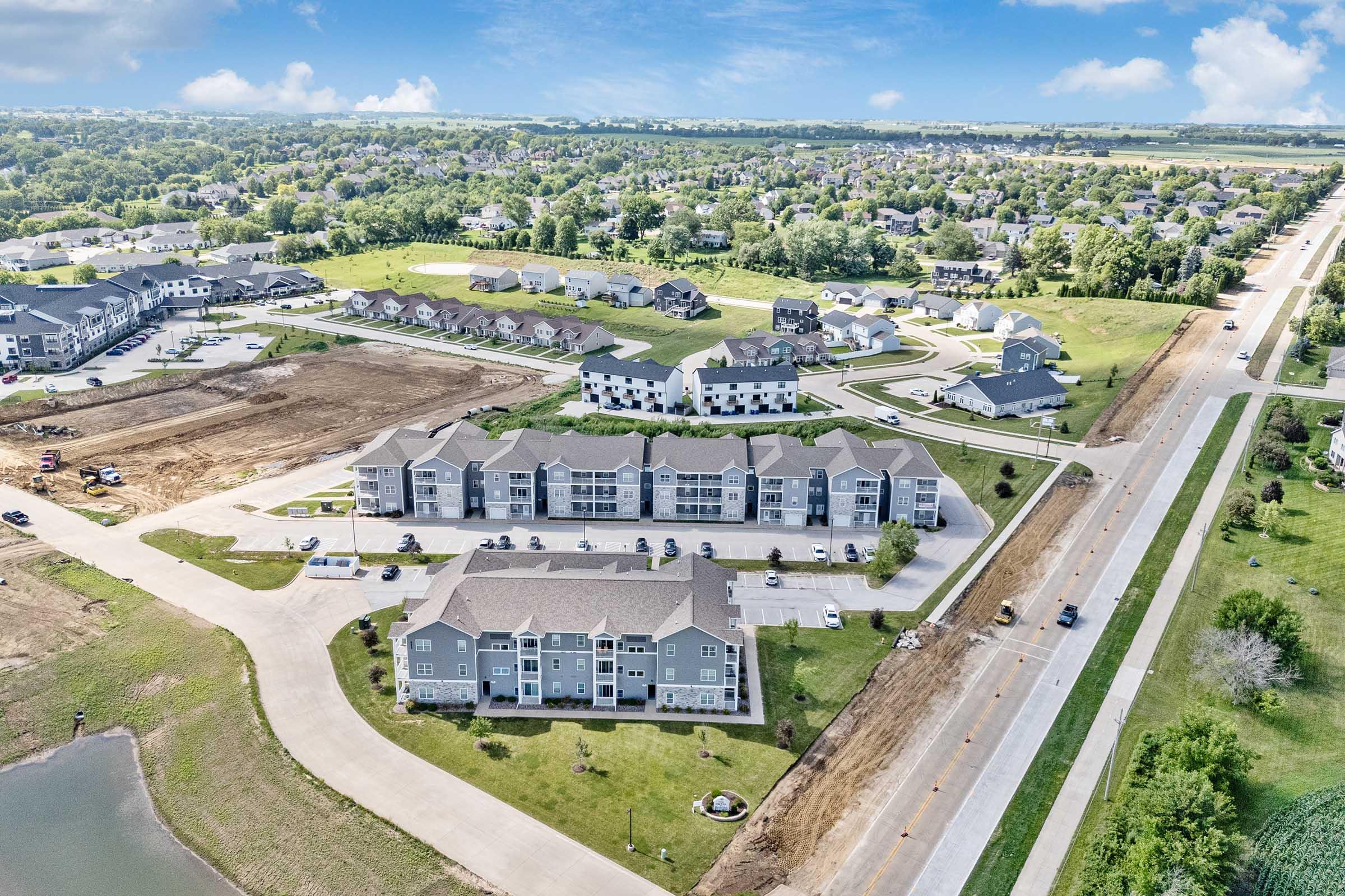 Aerial view of a suburban area featuring multiple apartment buildings, green lawn spaces, and nearby roads. Some construction activity is visible in the foreground, along with a lake or pond. The landscape is mostly green with residential neighborhoods in the distance.