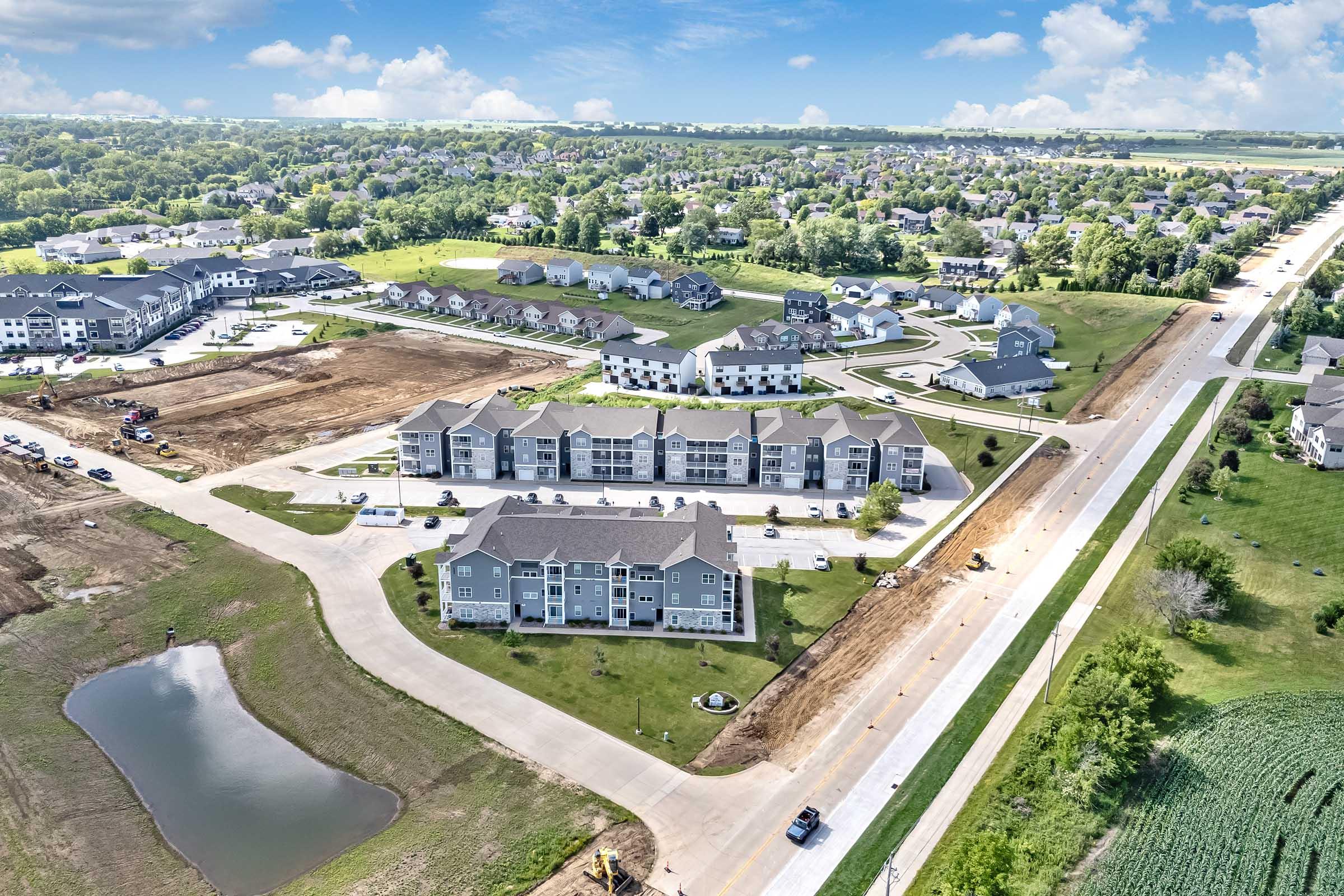 Aerial view of a residential area featuring multiple apartment buildings, green spaces, and a partially constructed road. Surrounding homes and open fields are visible, with a small pond in the foreground. The sky is clear and sunny, enhancing the vibrant colors of the landscape.