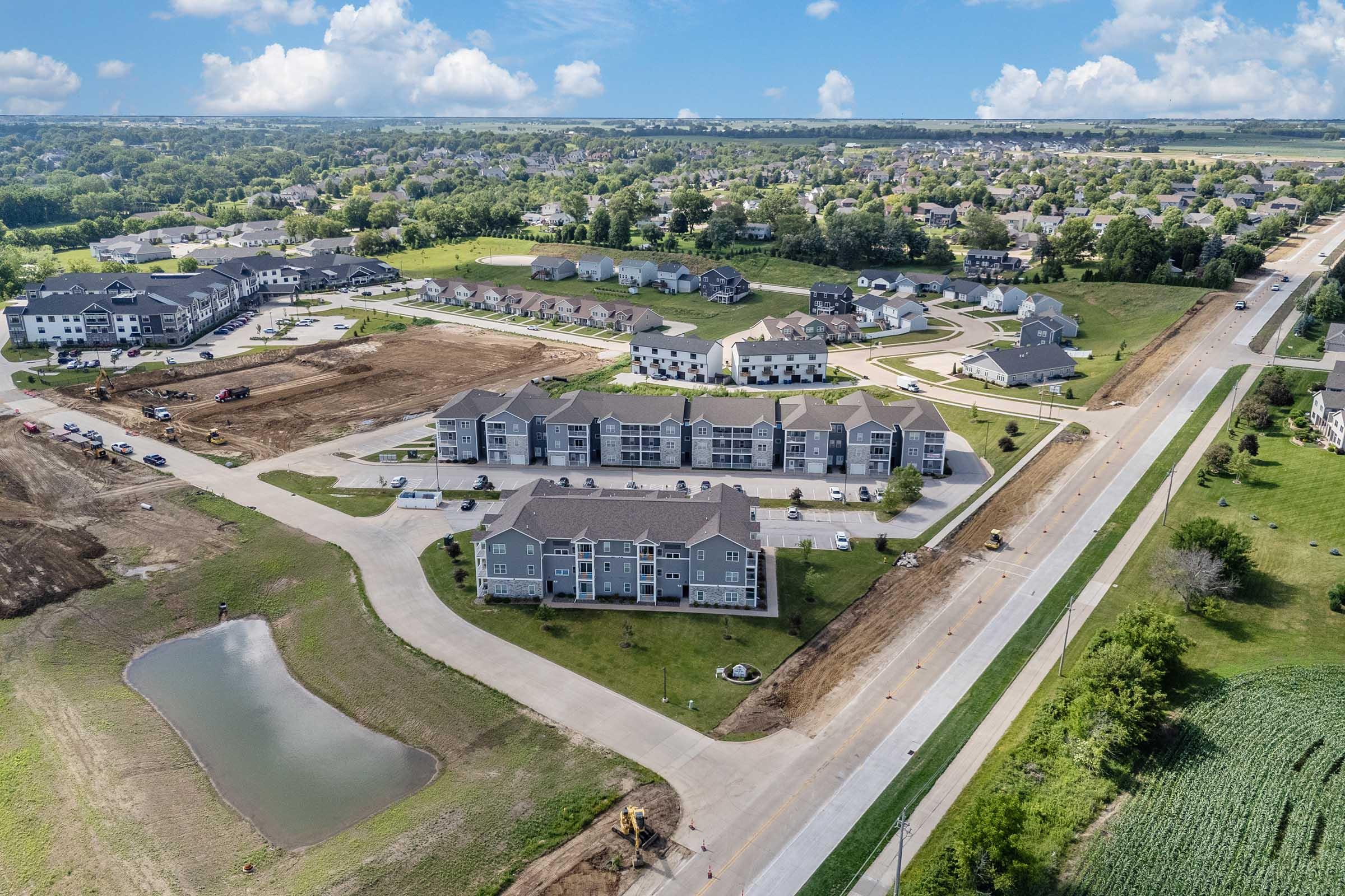 Aerial view of a residential area featuring a mix of multi-story apartment buildings and single-family homes. Roads and parking lots are visible, along with green spaces and a pond. The landscape shows a mix of developed and undeveloped land, with a blue sky and scattered clouds in the background.
