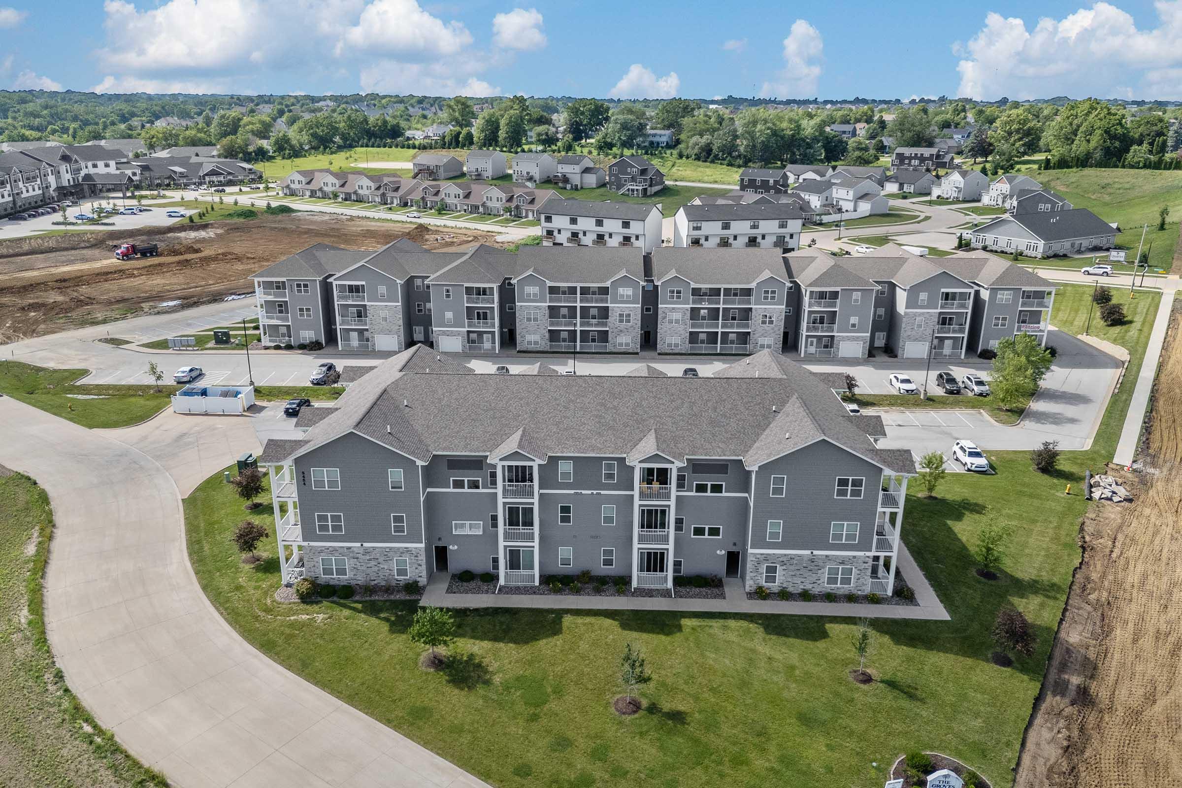 Aerial view of a modern apartment complex with gray siding and landscaped grounds. Surrounding area includes additional buildings and open space, with partially cleared land in the background. Bright blue sky with some clouds enhances the overall scenery.