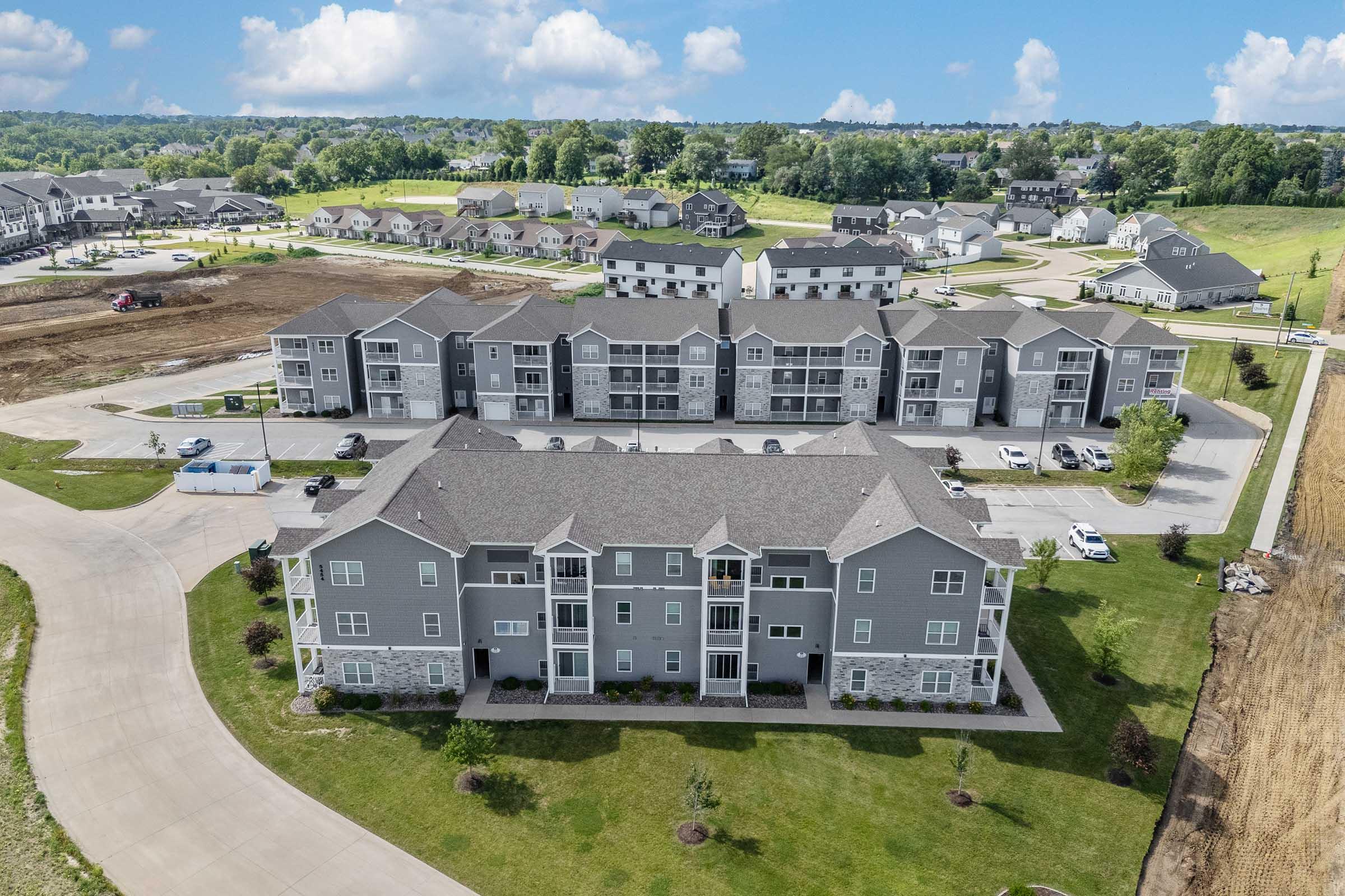 Aerial view of a modern apartment complex surrounded by landscaped grounds. The building features multiple stories with balconies, and is situated near residential homes and open fields. The sky is partly cloudy, and there are paved roads and parked vehicles in the vicinity.