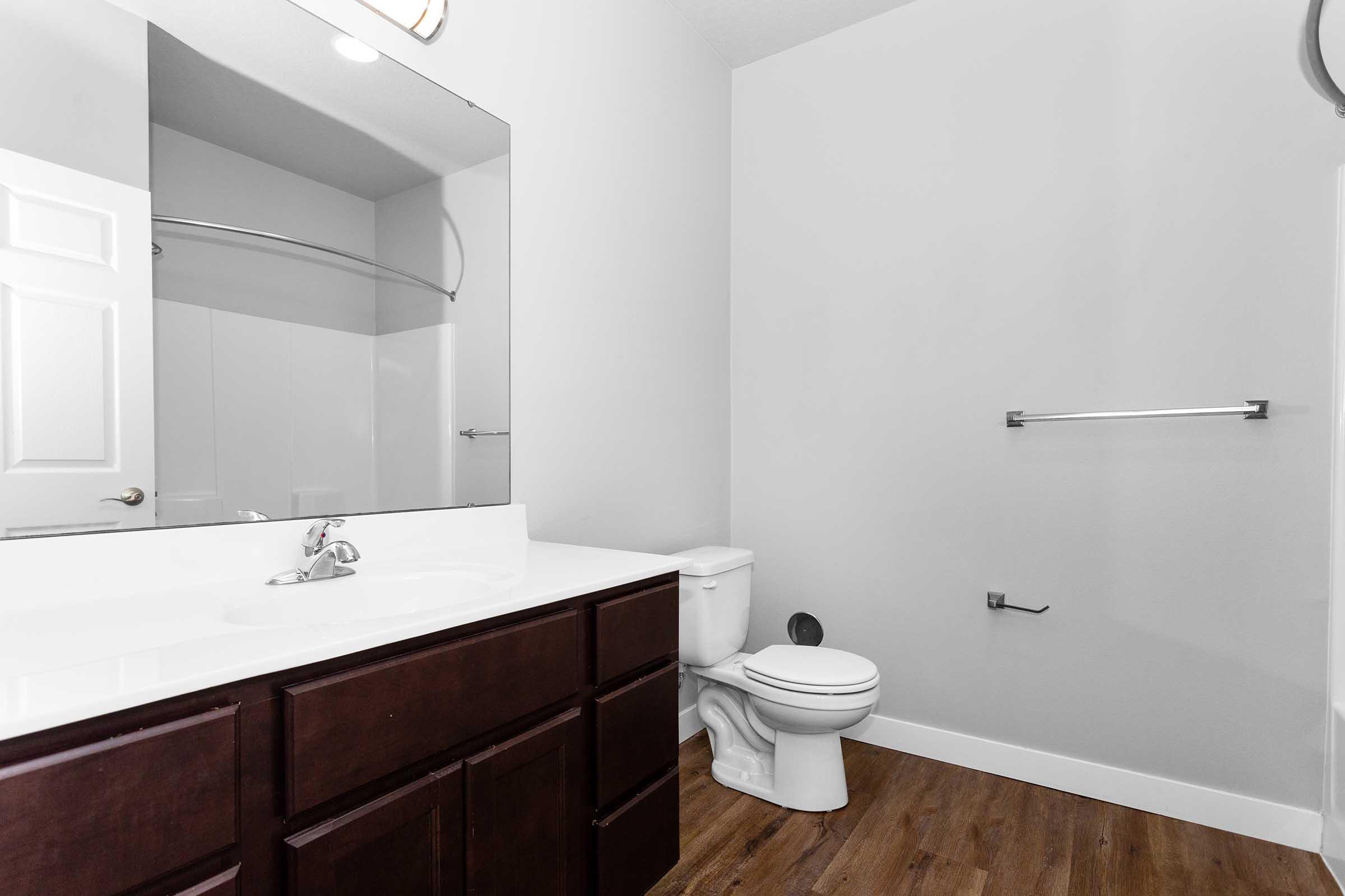 A modern bathroom featuring a large mirror above a white sink with brown cabinets underneath. A toilet is positioned to the right, and a towel bar is mounted on the wall. The walls are painted light gray, with a light wood floor giving a clean, minimalist appearance.