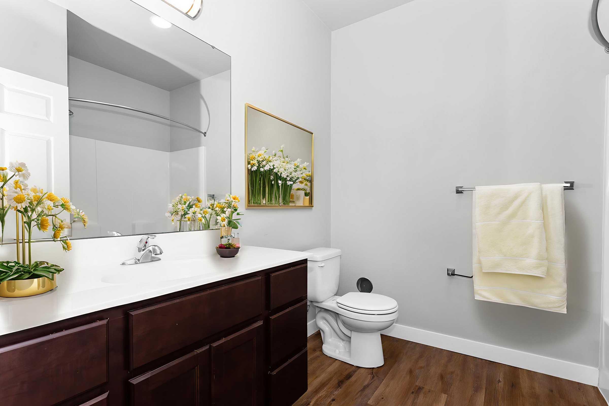 A bright, modern bathroom featuring a large mirror above a white sink with a dark wood vanity. A toilet is positioned beside the sink. A towel rack holds a light yellow towel, and a potted plant with yellow flowers adds a touch of color. The walls are painted light gray, and the flooring is wood-like.