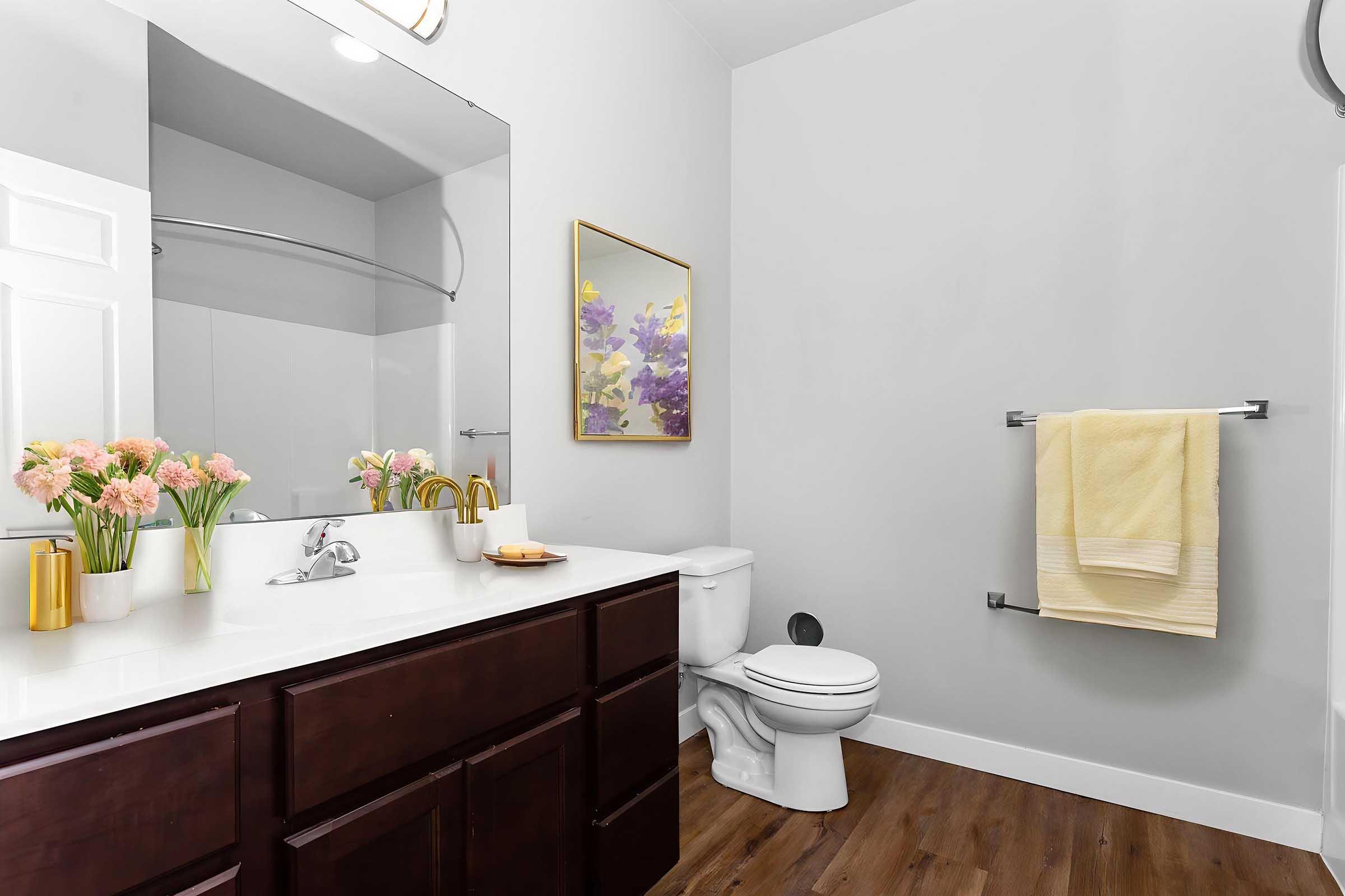 A modern bathroom featuring a white countertop with a vase of pink flowers, a large mirror, and an elegant wall painting. The space includes a white toilet, a towel rack with a yellow towel, and a glass shower enclosure, all set against light gray walls and wooden flooring.