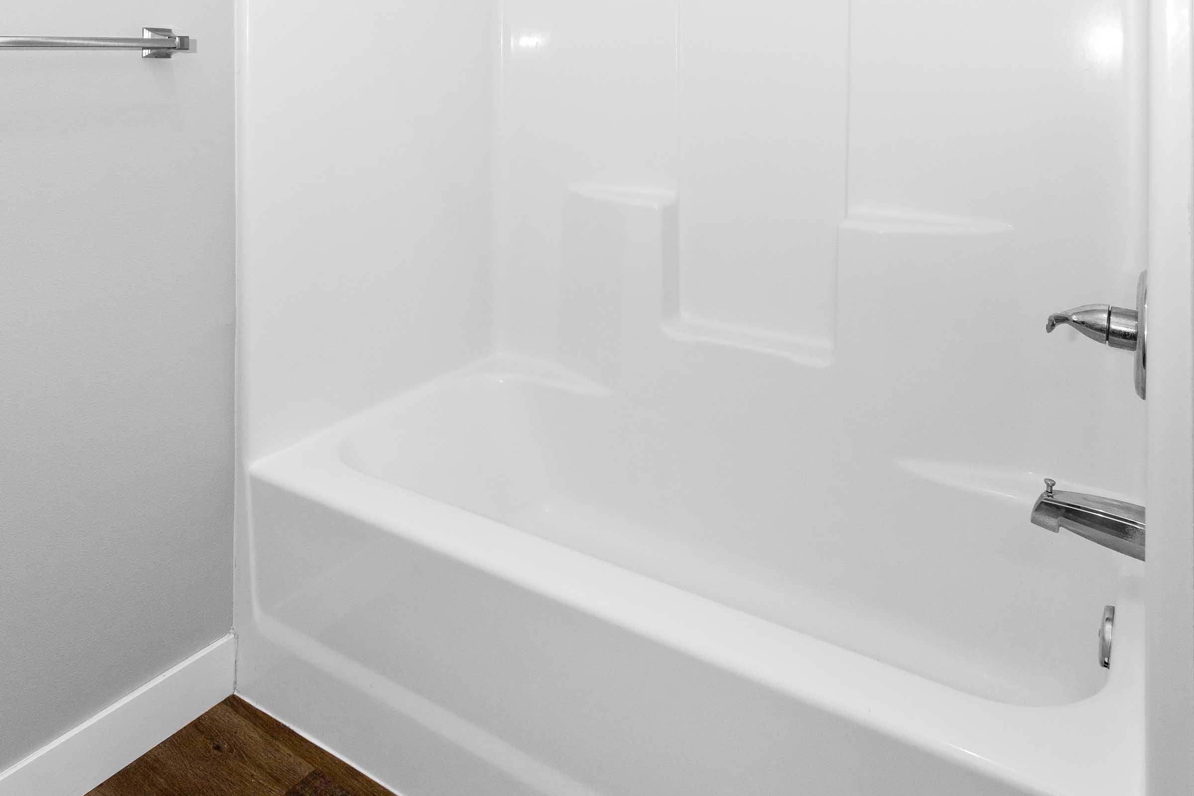 A clean, modern bathroom featuring a white bathtub with a shower fixture. The walls are smooth and white, and there is a chrome towel bar attached to the wall. The floor is wooden, adding a touch of warmth to the minimalist design.