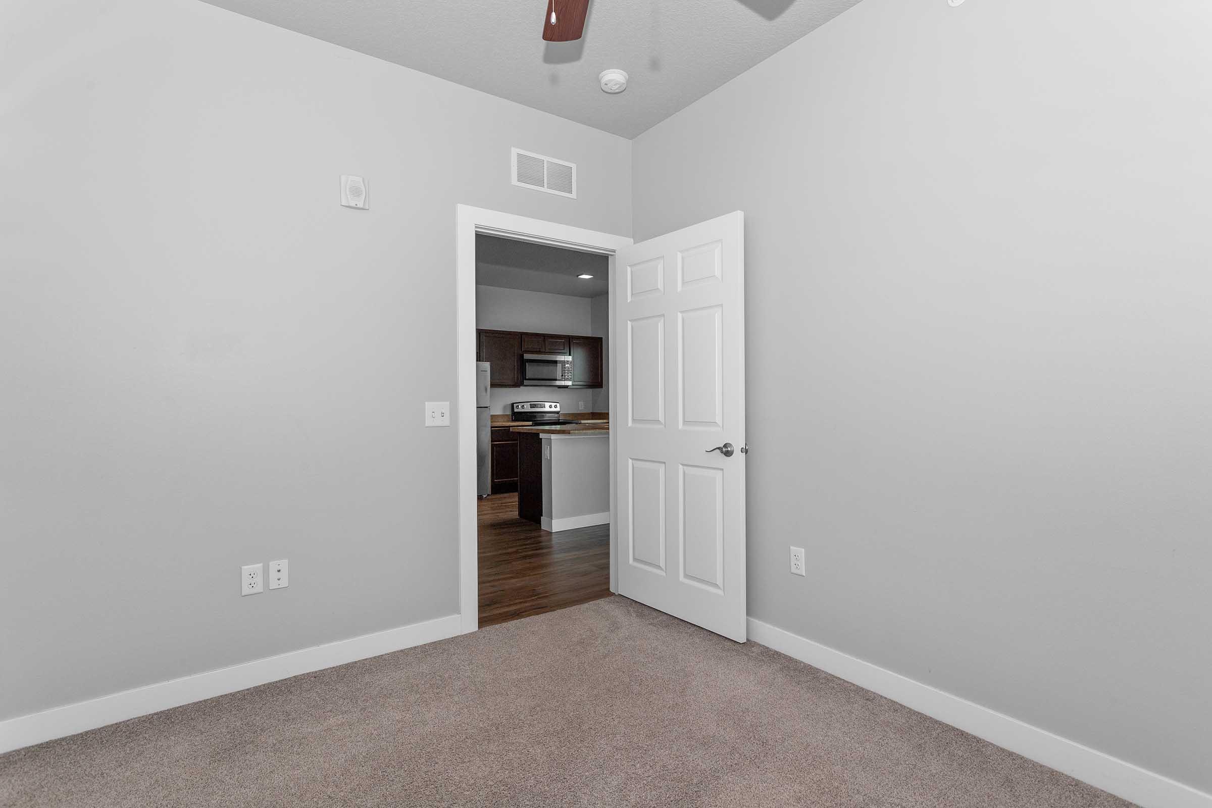 A well-lit room with light gray walls and carpeted flooring. The focal point is an open white door leading to an adjacent area, likely a kitchen, as indicated by a glimpse of cabinetry and appliances. A ceiling fan is visible, and there are no furnishings in the room.