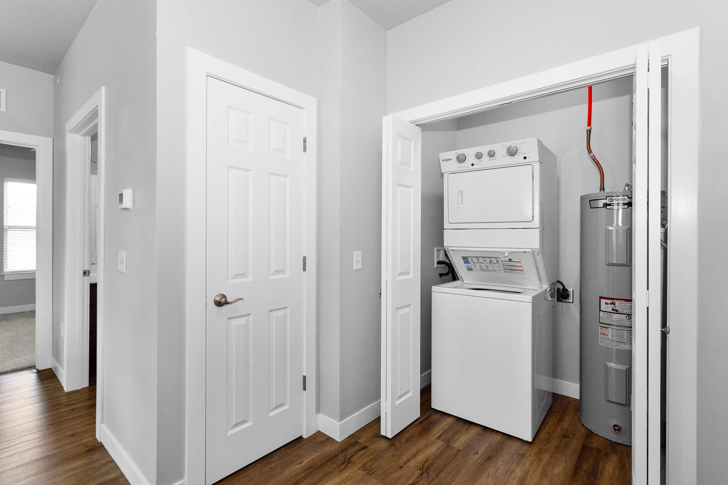A laundry area featuring a stacked washer and dryer in a closet, alongside a water heater. The space has light gray walls and wooden flooring, with a door leading to another room visible in the background.