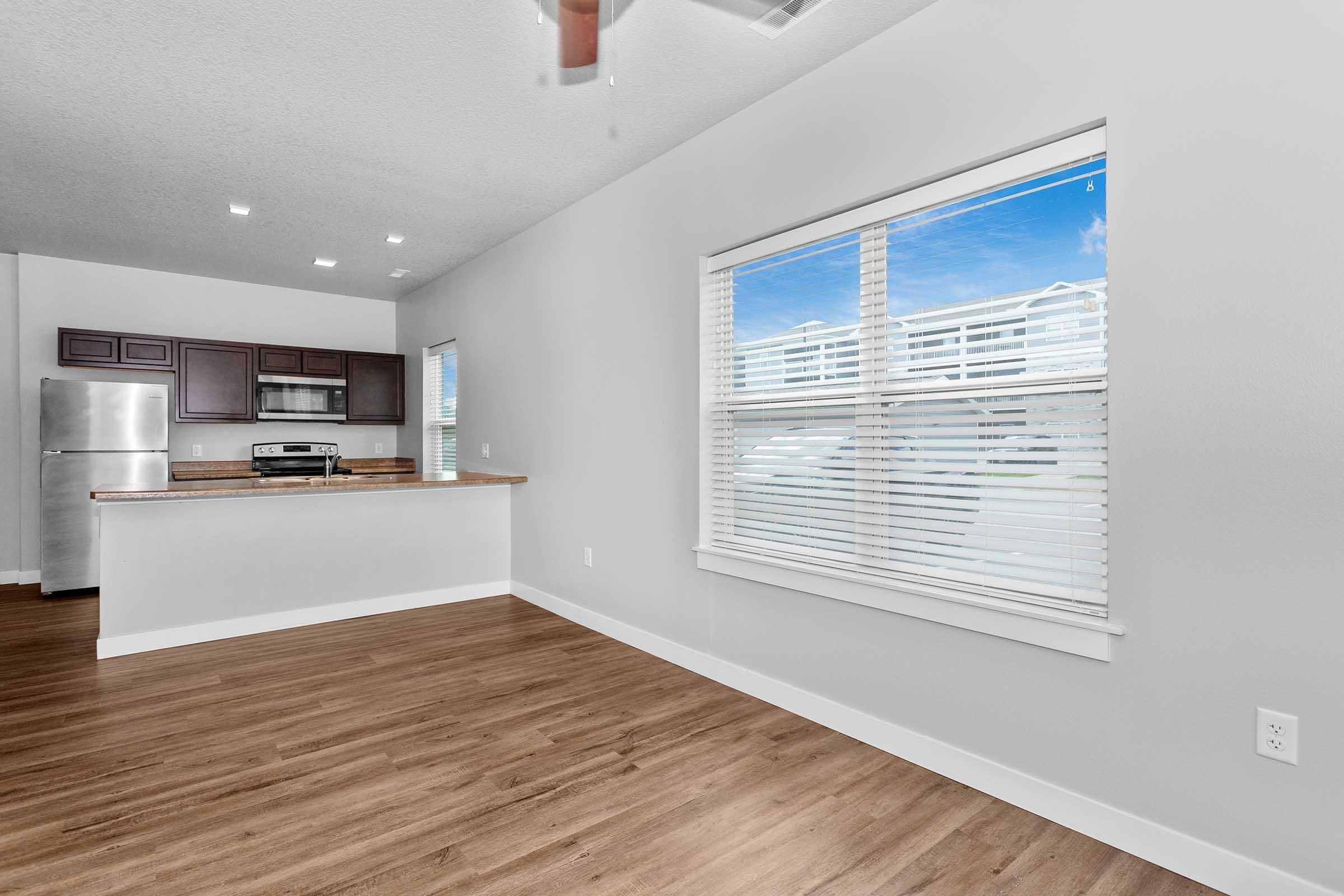 A modern, open-concept living space featuring a hardwood floor, a large window with white blinds letting in natural light, and a kitchen area with dark wood cabinets and stainless steel appliances in the background. The walls are light-colored, creating a bright and airy atmosphere.