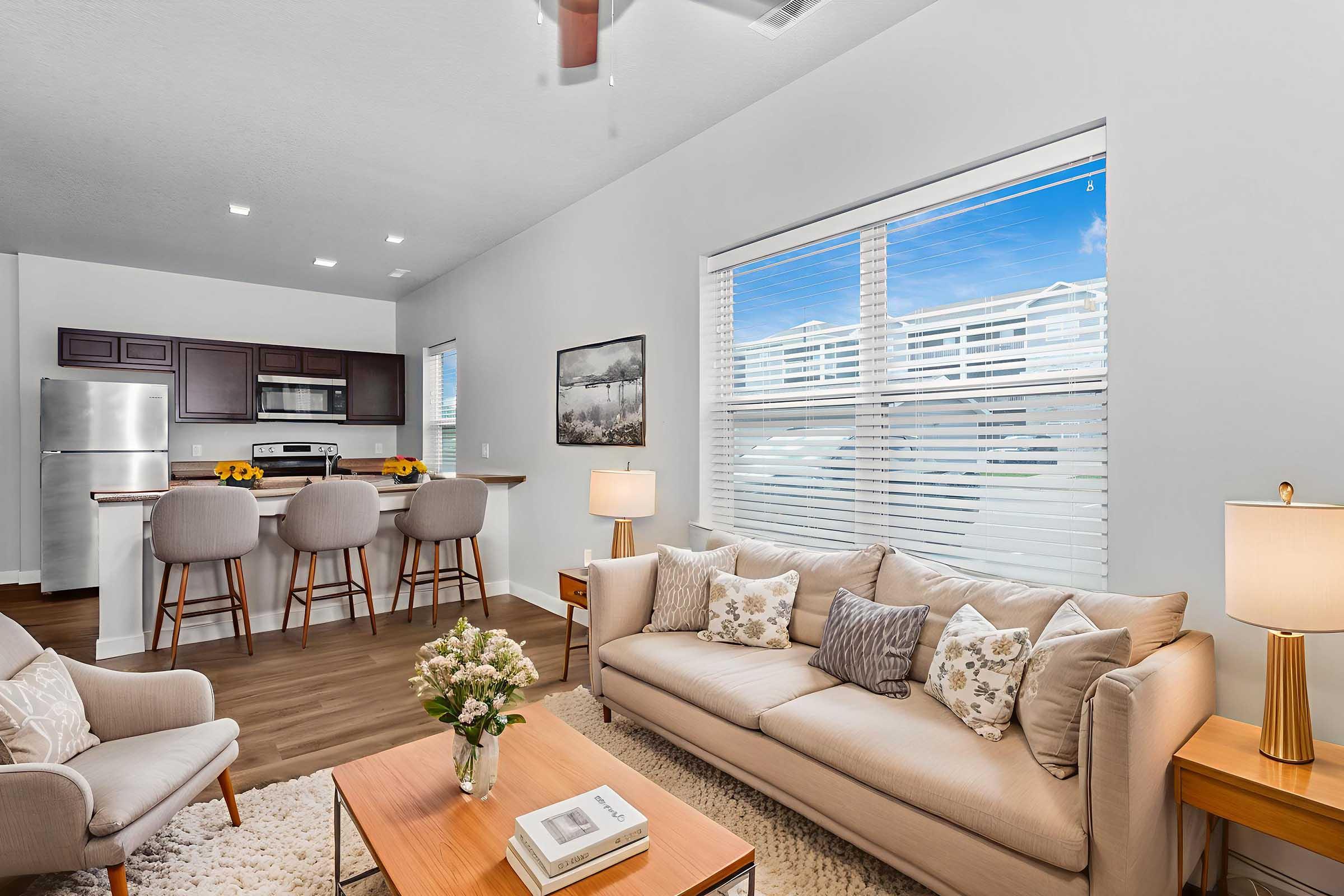 A modern living room featuring a beige sofa adorned with decorative pillows, a wooden coffee table with a vase of flowers, and a cozy rug. In the background, a bright kitchen with stainless steel appliances and barstools is visible. Large windows provide natural light and a view of the outdoors.