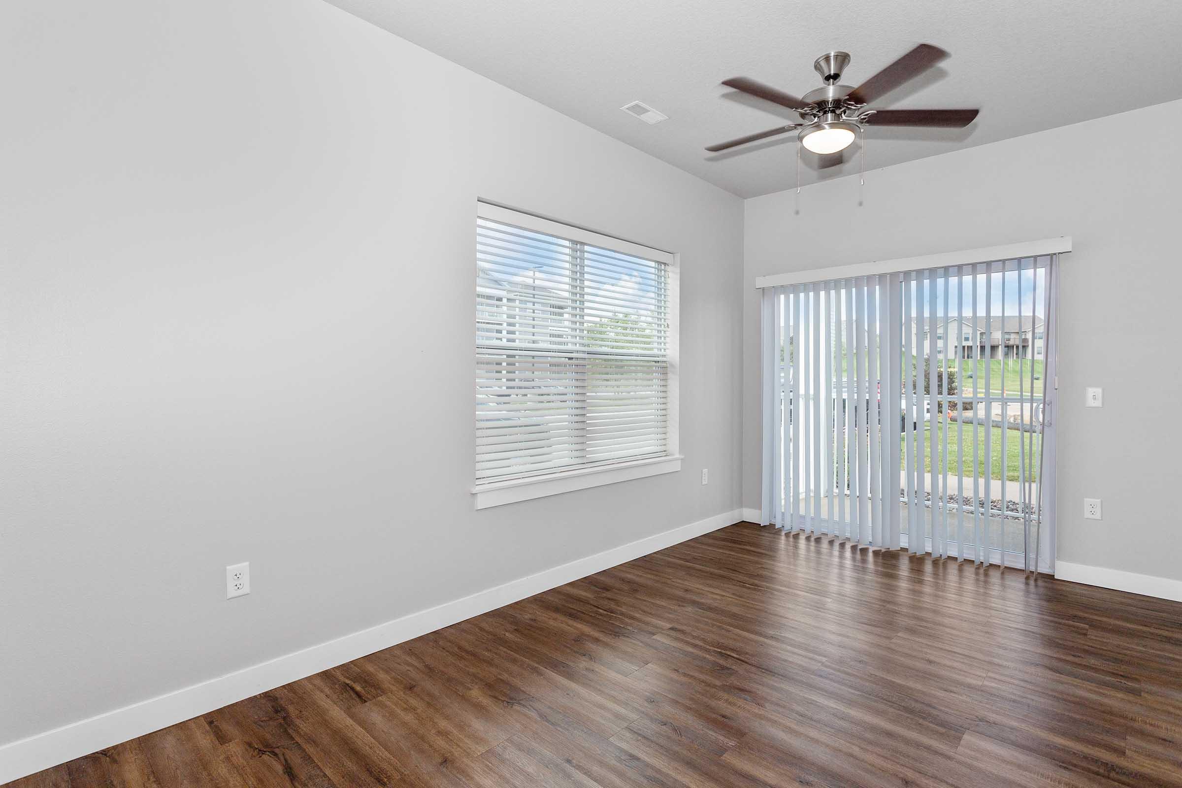 A well-lit room with light gray walls and a large window featuring white blinds. The floor is finished in a warm wood tone, and a ceiling fan is installed. A sliding glass door with vertical blinds leads to an outdoor space. The room appears spacious and invites natural light.