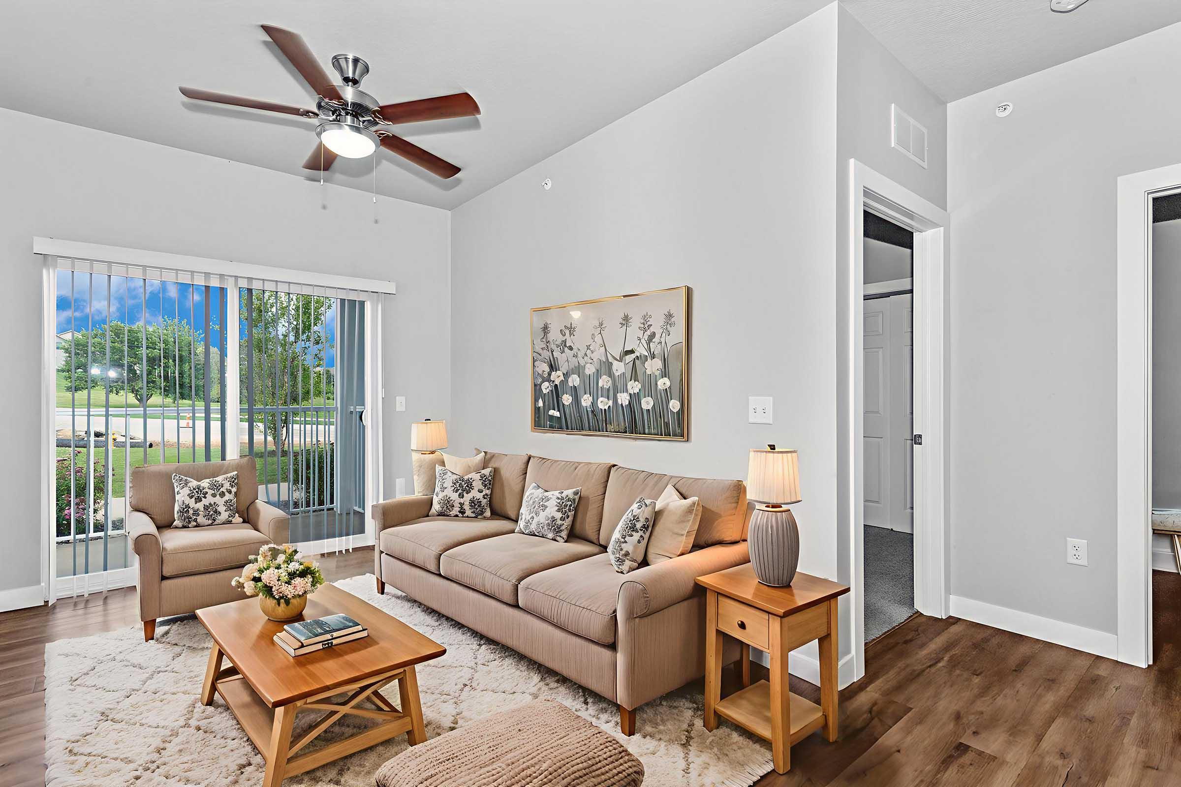 Cozy living room featuring a beige sofa with accent pillows, a wooden coffee table with a floral arrangement, and a beige armchair. A ceiling fan hangs above, and large windows provide natural light and views of greenery outside. The decor includes a framed wall art piece and warm wooden flooring.