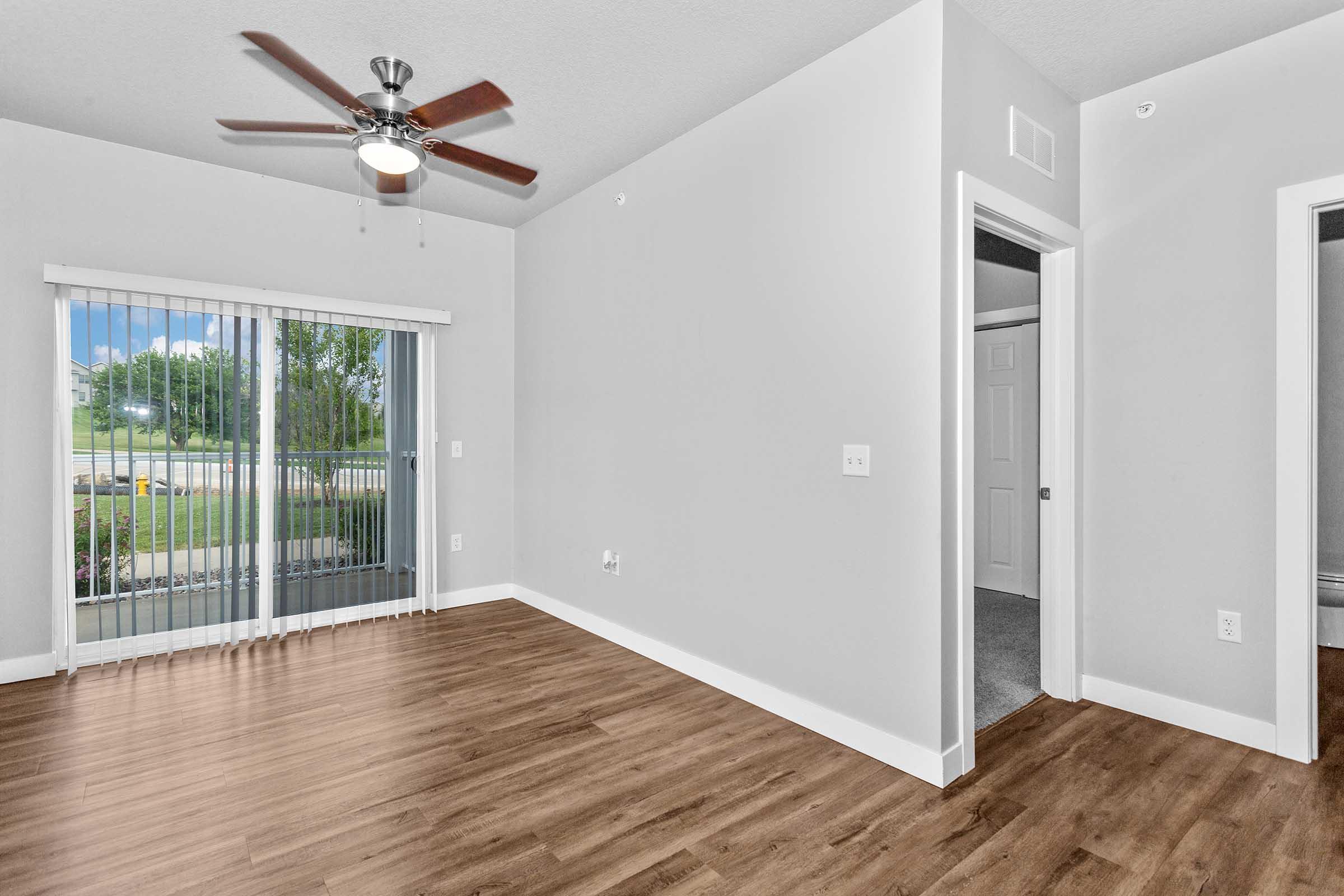 Spacious interior of a light-colored room featuring a ceiling fan, large window with blinds, and a sliding glass door leading to a patio. The floor is wood-like, and there are white walls. A doorway leads to another room, showcasing a modern and inviting living space.