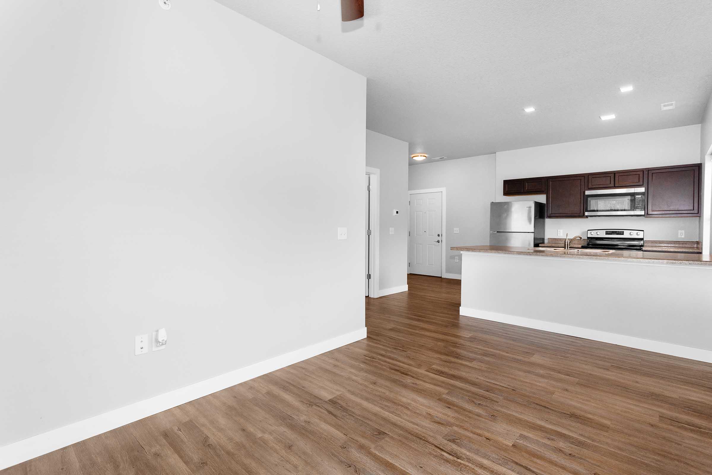 A spacious, modern kitchen and living area with light wood flooring and gray walls. The kitchen features dark wooden cabinets, stainless steel appliances, and a breakfast bar. There is a doorway leading to a hallway, and overhead lighting brightens the open space.