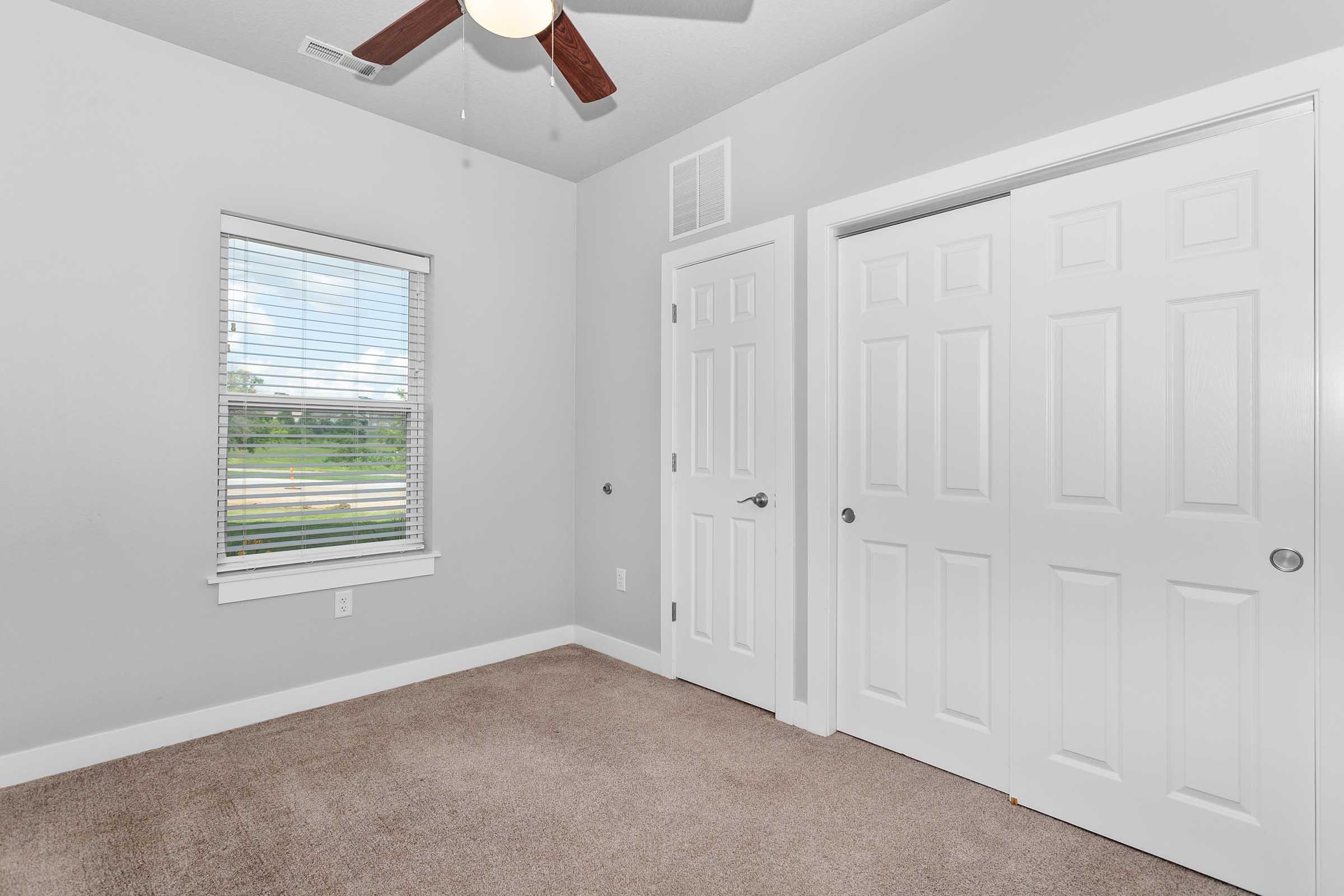 A small, empty room with light gray walls and carpet flooring. It features a window with blinds, allowing natural light, and a ceiling fan. There are two white closet doors on one side, and the room appears tidy and well-maintained, suggesting a modern and inviting atmosphere.