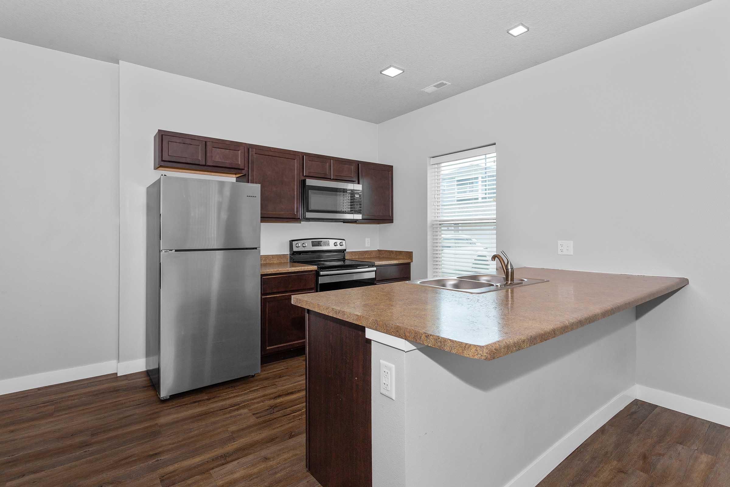 A modern kitchen featuring dark wood cabinets, stainless steel appliances including a refrigerator and oven, and a single-basin sink. The countertop is spacious, and there is a window providing natural light. The flooring is a warm-toned laminate.
