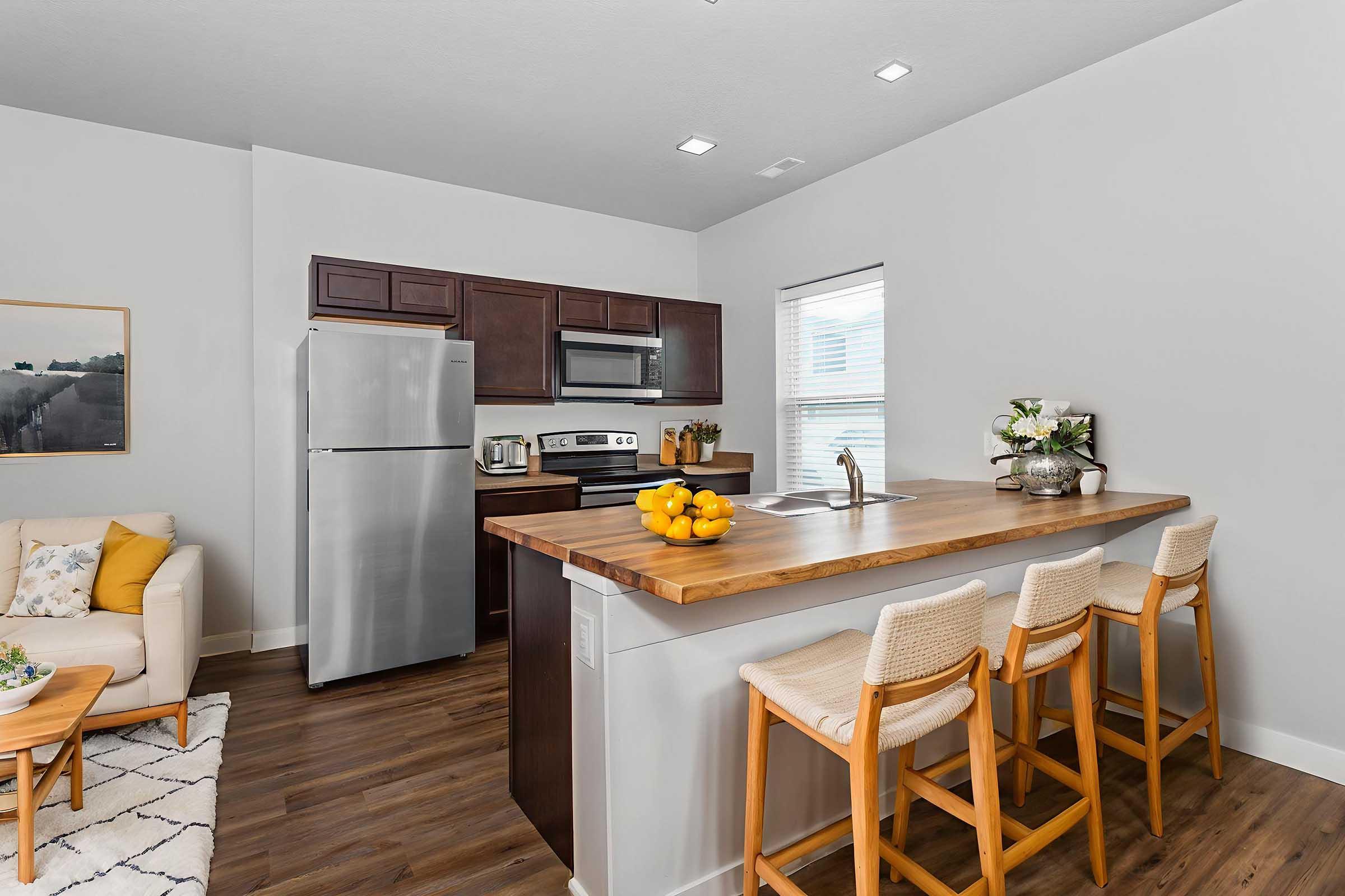 Modern kitchen with dark wood cabinets and stainless steel appliances. A spacious island with a wooden countertop and four bar stools. Countertop displays a bowl of oranges and a few decorative items. Light-colored sofa and a cozy rug are visible in the connecting living area, enhancing the inviting atmosphere.