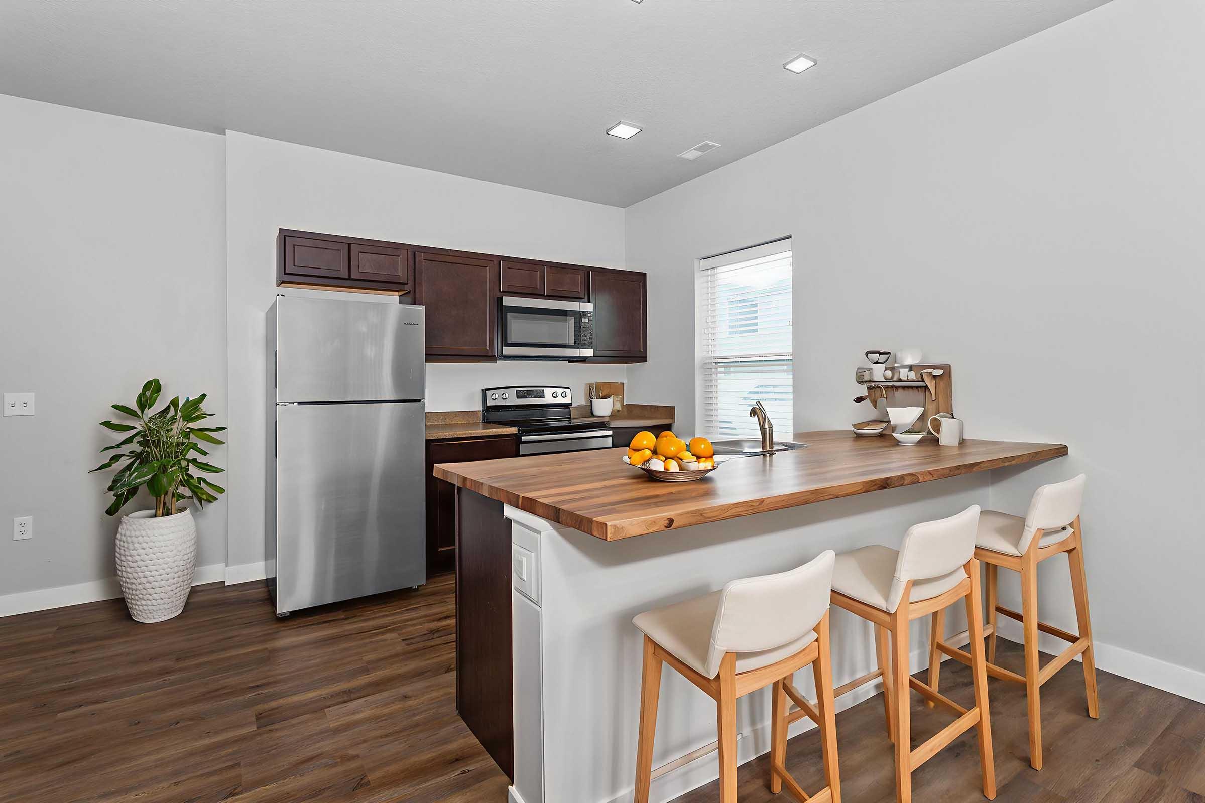 Modern kitchen with stainless steel appliances, including a refrigerator and oven. A wooden bar area with white stools is in the foreground, adorned with a bowl of oranges. A green potted plant is placed nearby, and there is natural light coming through a window, enhancing the bright and inviting atmosphere.