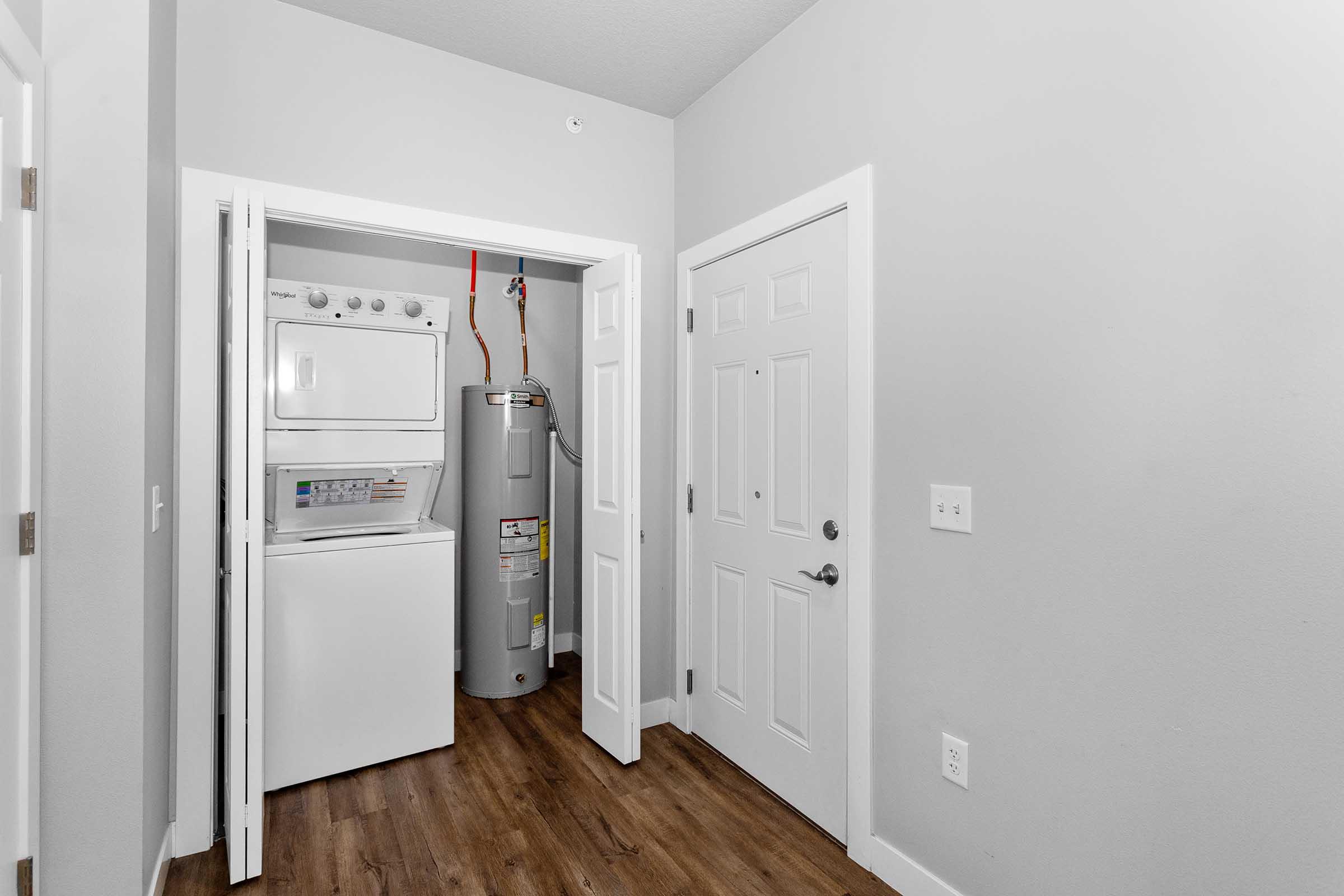 A small laundry room featuring a stacked washer and dryer unit and a water heater. The room has light gray walls and a wood-like floor. There is a door leading outside and a closet on one side, creating a functional and compact space.