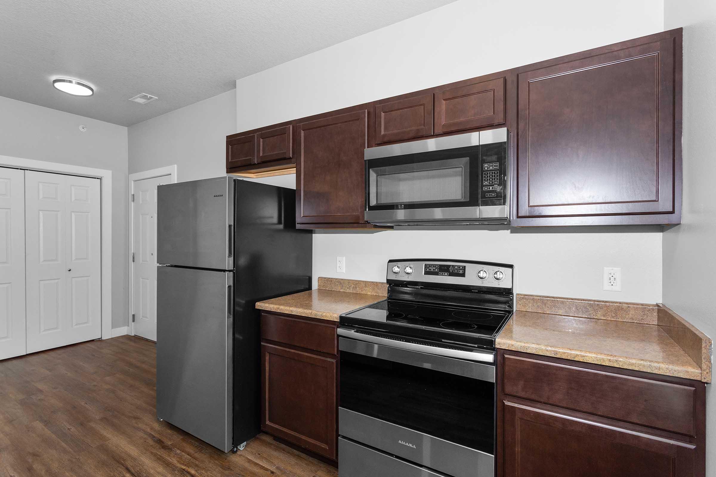 Modern kitchen featuring dark wood cabinets, a stainless steel refrigerator, a microwave above an electric stove, and a countertop. The flooring is a warm wood tone, and there are light-colored walls. A door leading to another room is visible in the background.