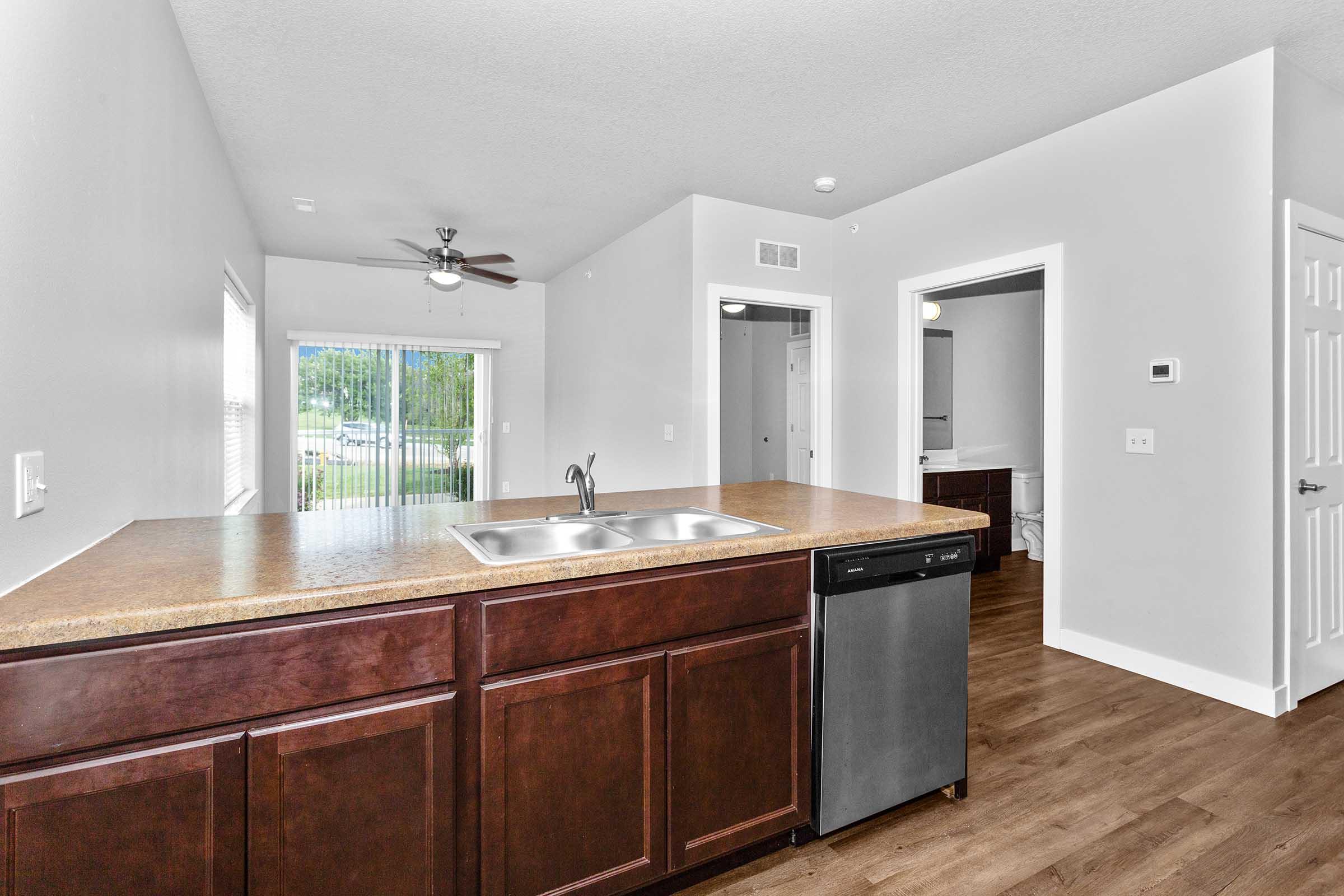 A modern kitchen view featuring a brown wood counter with a sink and dishwasher, light gray walls, and a ceiling fan. Natural light enters from a nearby window, and an open doorway leads to another room, suggesting a spacious and bright living area.