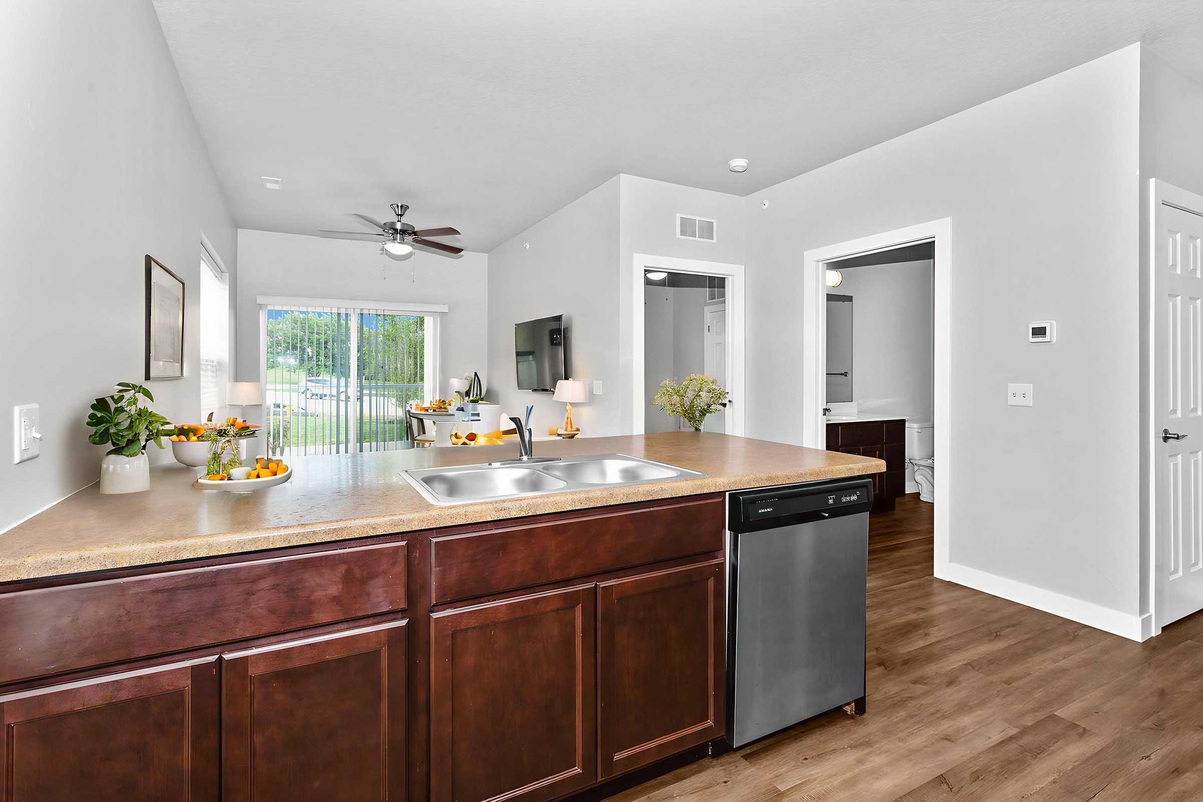 A modern kitchen with dark wood cabinetry, a double sink, and stainless steel dishwasher. Fresh fruit is displayed on the countertop. In the background, a bright living area features sliding glass doors leading to an outdoor space. Light-colored walls and wooden flooring enhance the spacious feel.