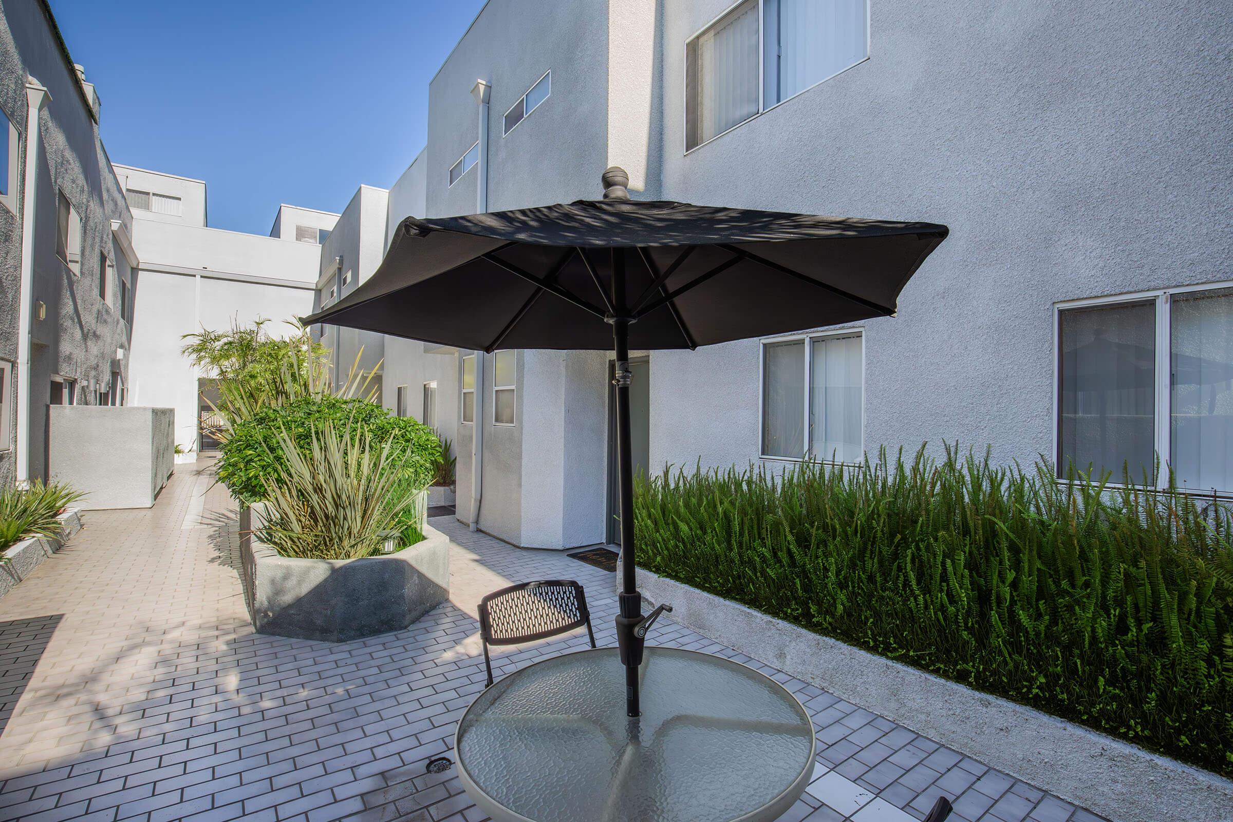 A small outdoor area featuring a black umbrella over a glass table. Surrounding the table are low shrubs and plants, with modern, gray buildings in the background and clear blue skies above. The scene conveys a serene and inviting atmosphere for relaxation.