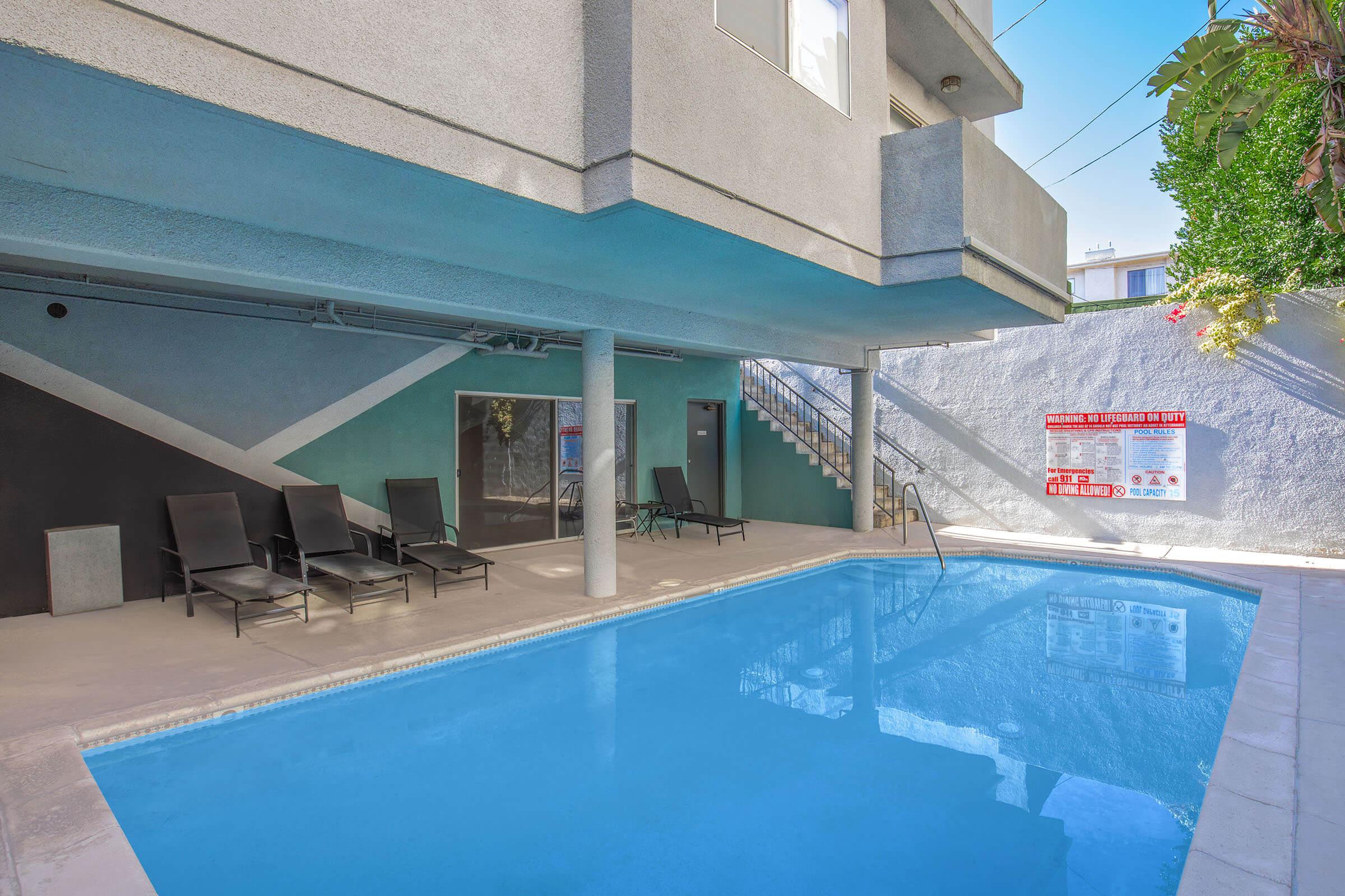 A modern swimming pool area with blue water surrounded by lounge chairs and a staircase. The walls feature geometric patterns, and there's a notice board displaying pool rules and information. Green plants are visible, adding a touch of nature to the space.