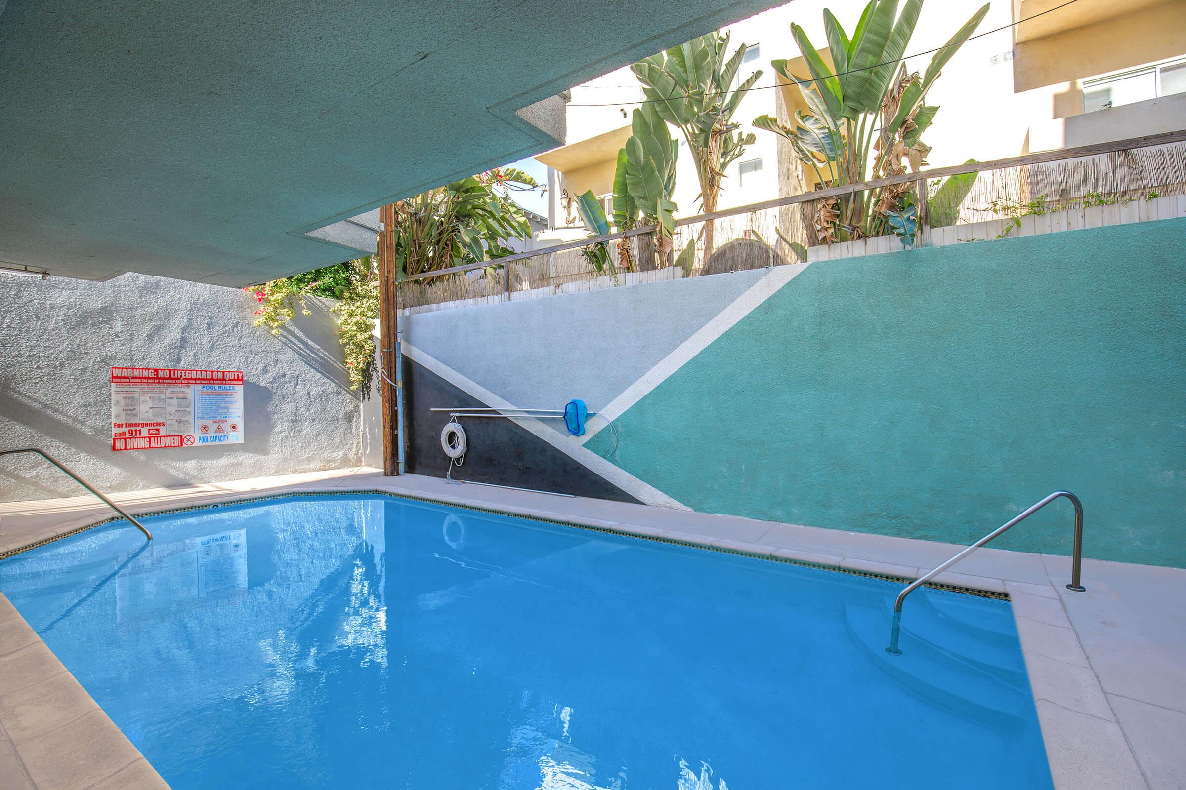 A serene pool area with clear blue water surrounded by a textured green wall and tropical plants. The space is partially shaded, with a lifebuoy mounted on the wall and a sign visible in the background. The setting offers a calm and inviting atmosphere.