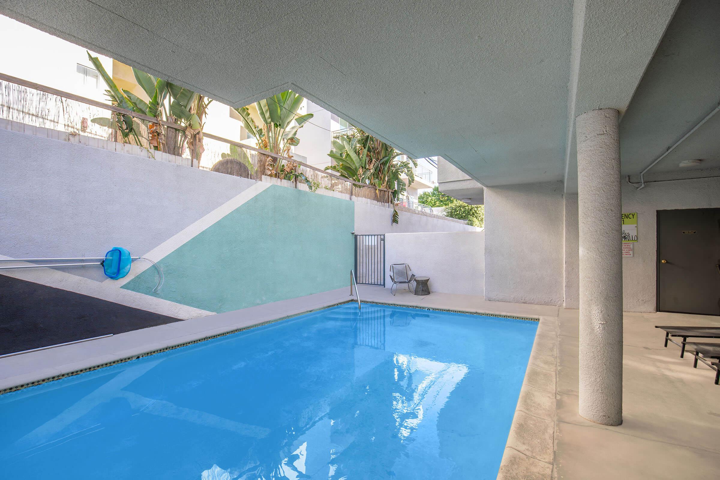 A clean, blue swimming pool surrounded by a concrete area and plants. The pool is situated under an overhang, with a lounge chair nearby. A painted wall features geometric shapes in muted colors. Sunlight brightens the space, creating a relaxing atmosphere.